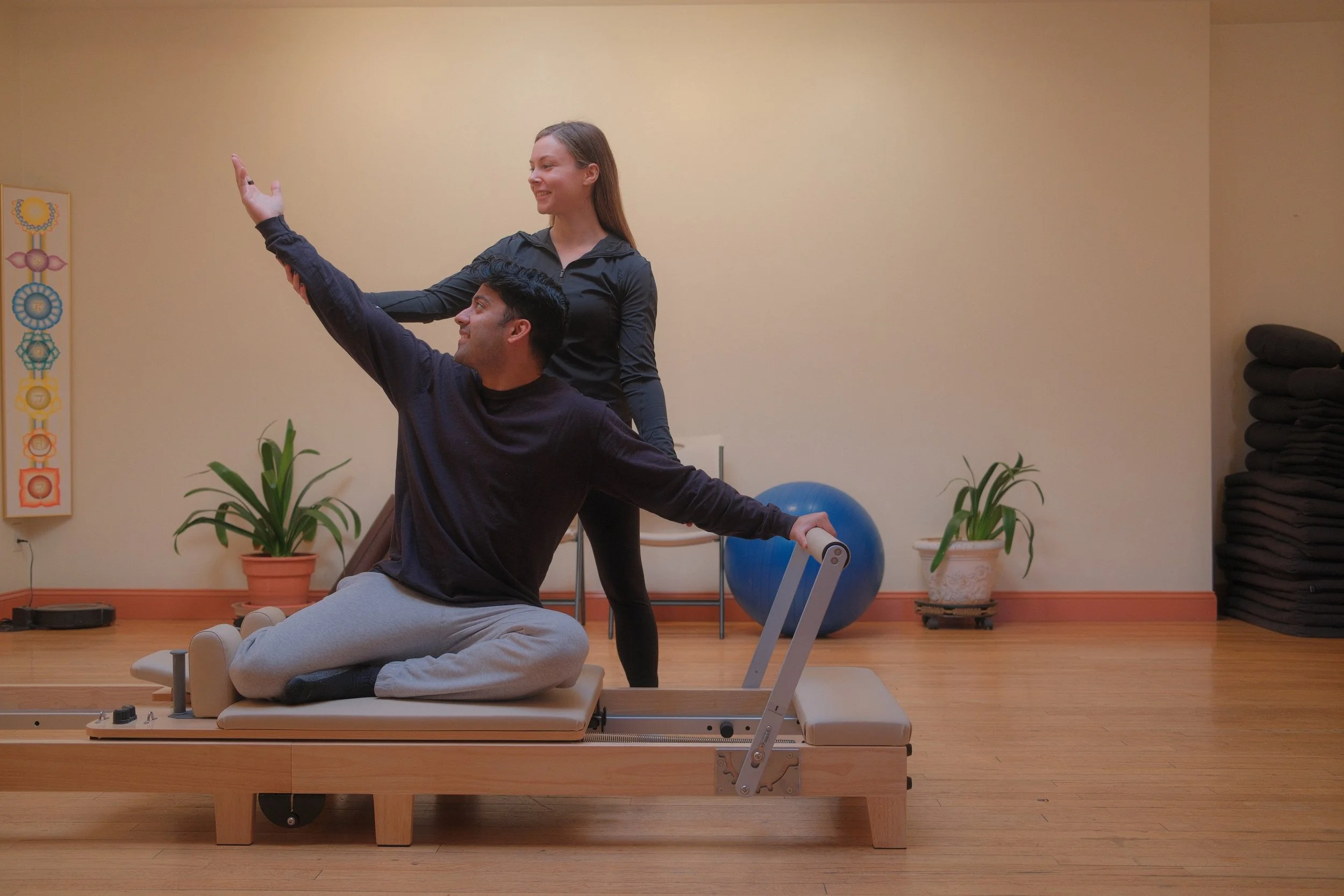 A physical therapist guides a man on a Pilates reformer machine during a therapy session in a rehab center, with exercise equipment and plant decorations in the background.