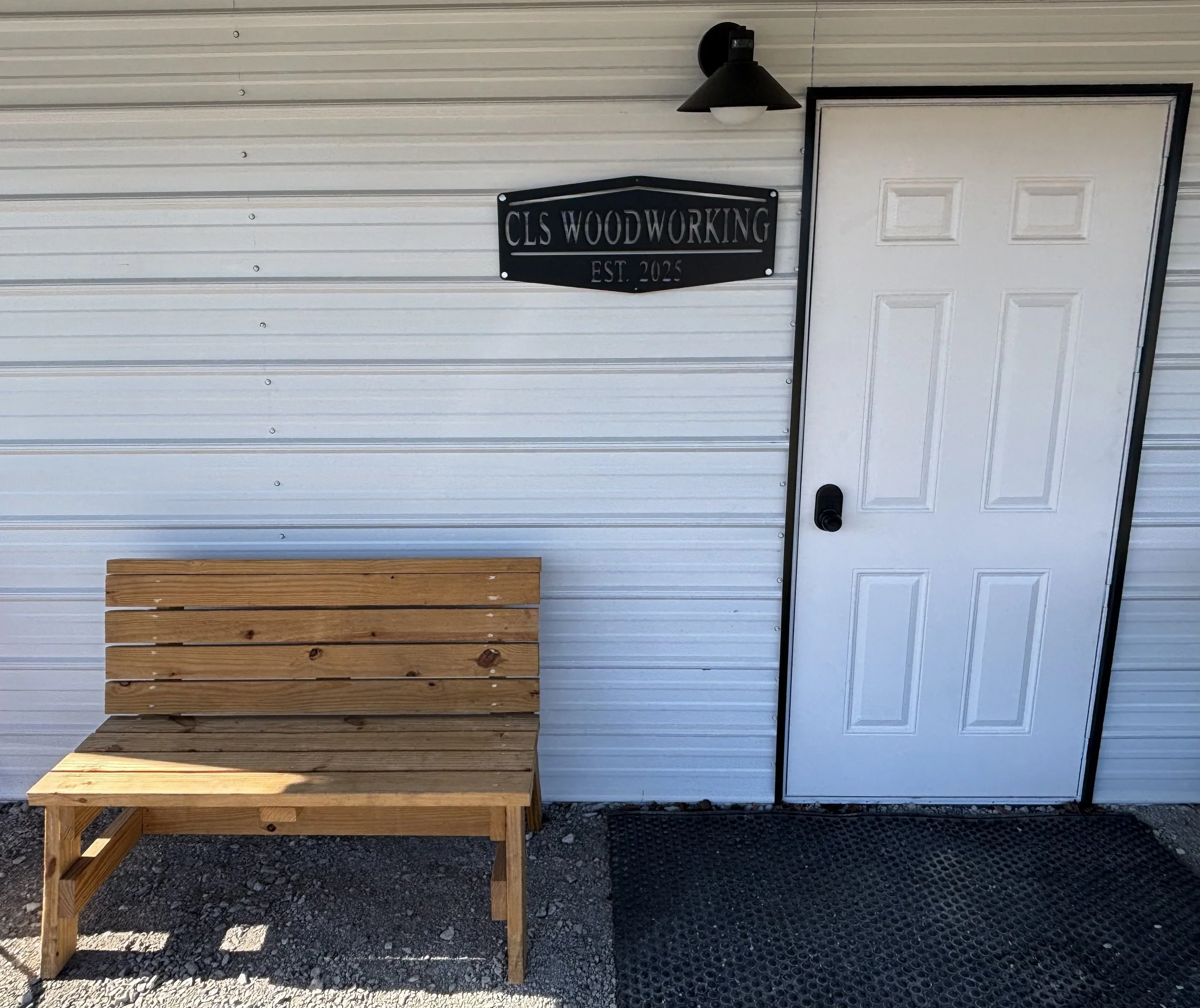 Wooden bench outside a building with a white door, black sign reading "CLS Woodworking EST. 2025," a black outdoor light above the sign, and a black doormat on the ground.