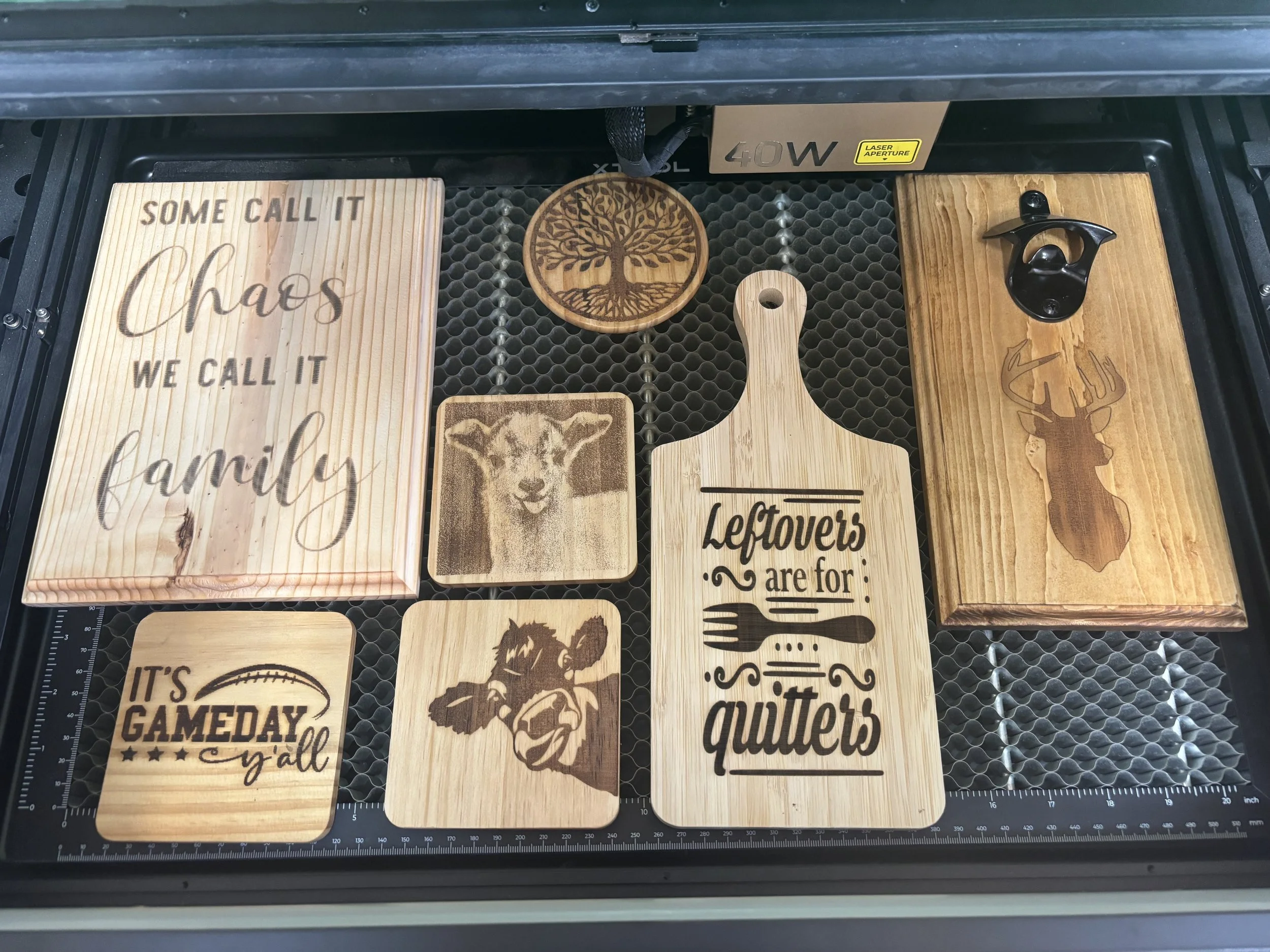 Collection of wooden engraved signs and coasters with various designs and quotes, including a tree, a lamb, a deer head, a dog face, and football-themed text.