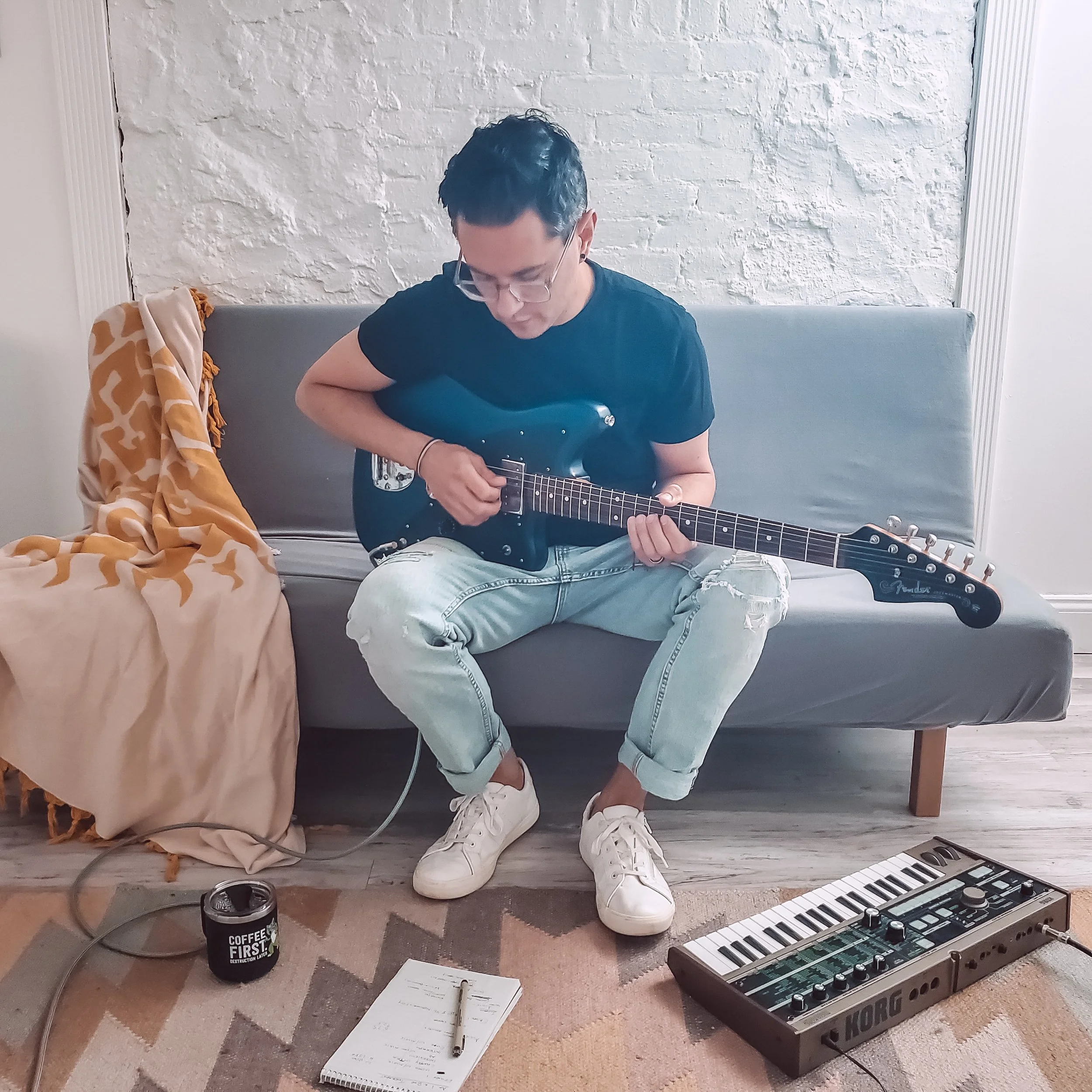 A person with short dark hair and glasses, wearing a black t-shirt and ripped jeans, sitting on a gray couch while playing an electric guitar. Music equipment including a keyboard, a notebook, a pen, a coffee cup, and an audio interface are on the floor nearby. The wall behind has white brick. There is a patterned blanket on the armrest of the couch.