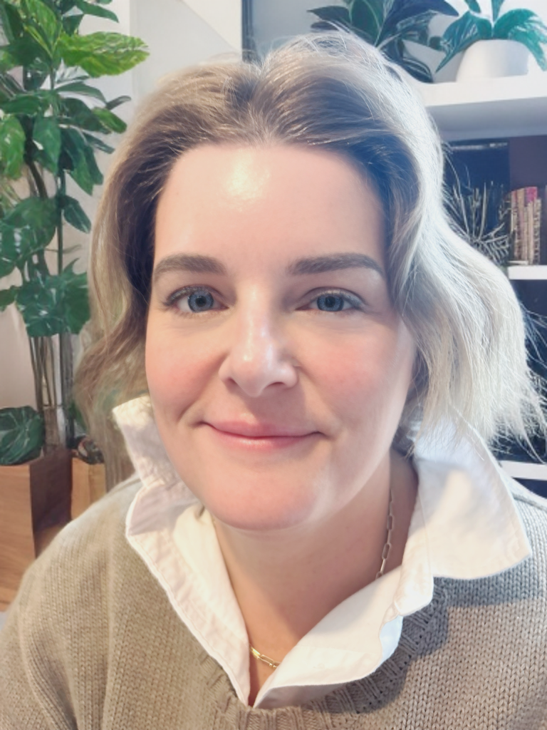 A woman with short wavy blonde hair, blue eyes, and fair skin, smiling in an indoor setting with plants and books in the background.