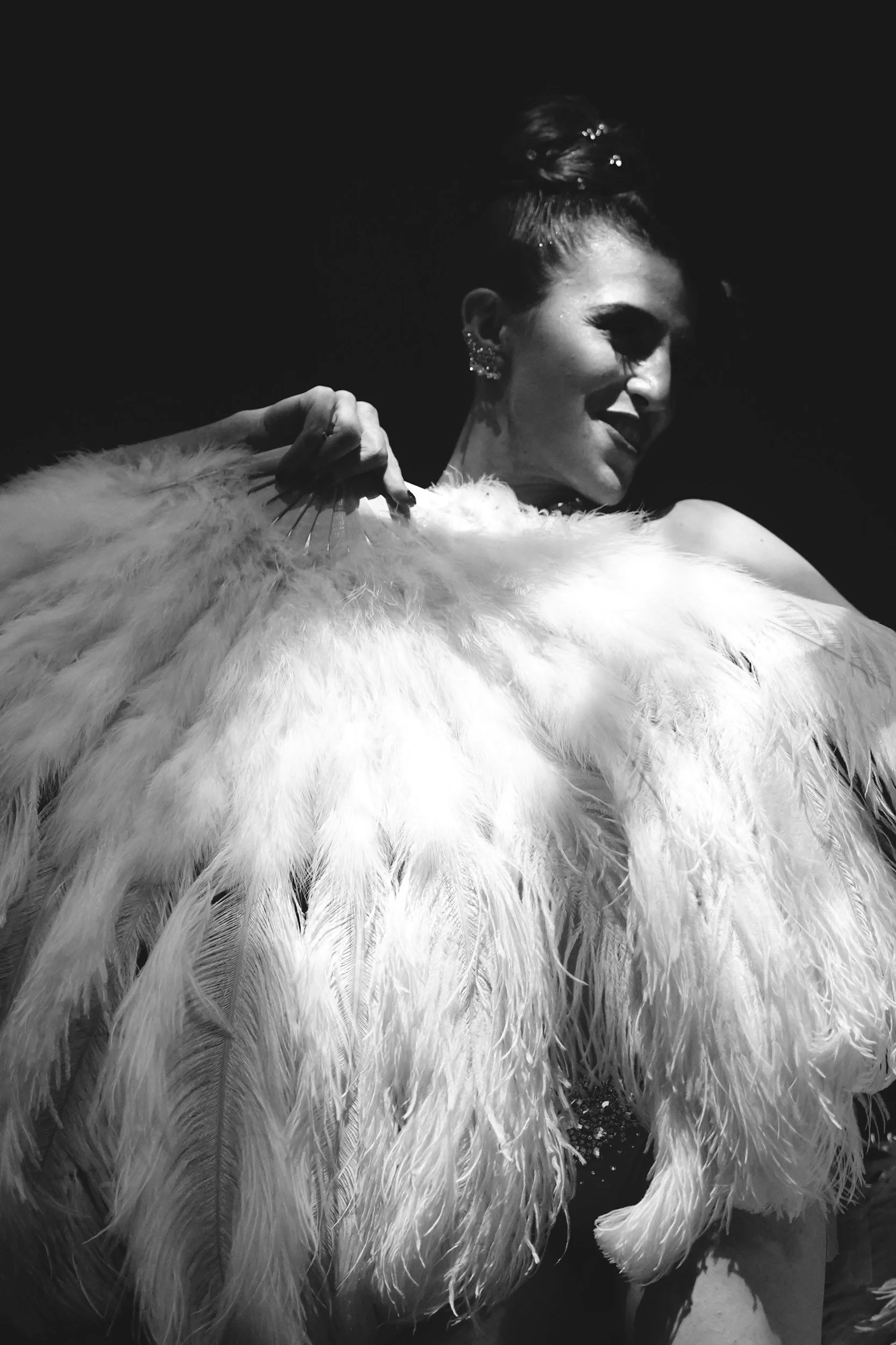 A woman with an elegant hairstyle and earrings is holding a large feathered fan or costume piece. The image is in black and white, with high contrast highlighting her smile and the feather details.