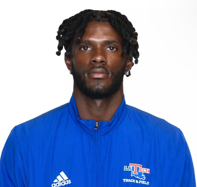 A man with dark skin, black curly hair, and a beard, wearing a blue athletic jacket with a zipper, Adidas logo, and LA Tech Track & Field emblem, against a plain white background.
