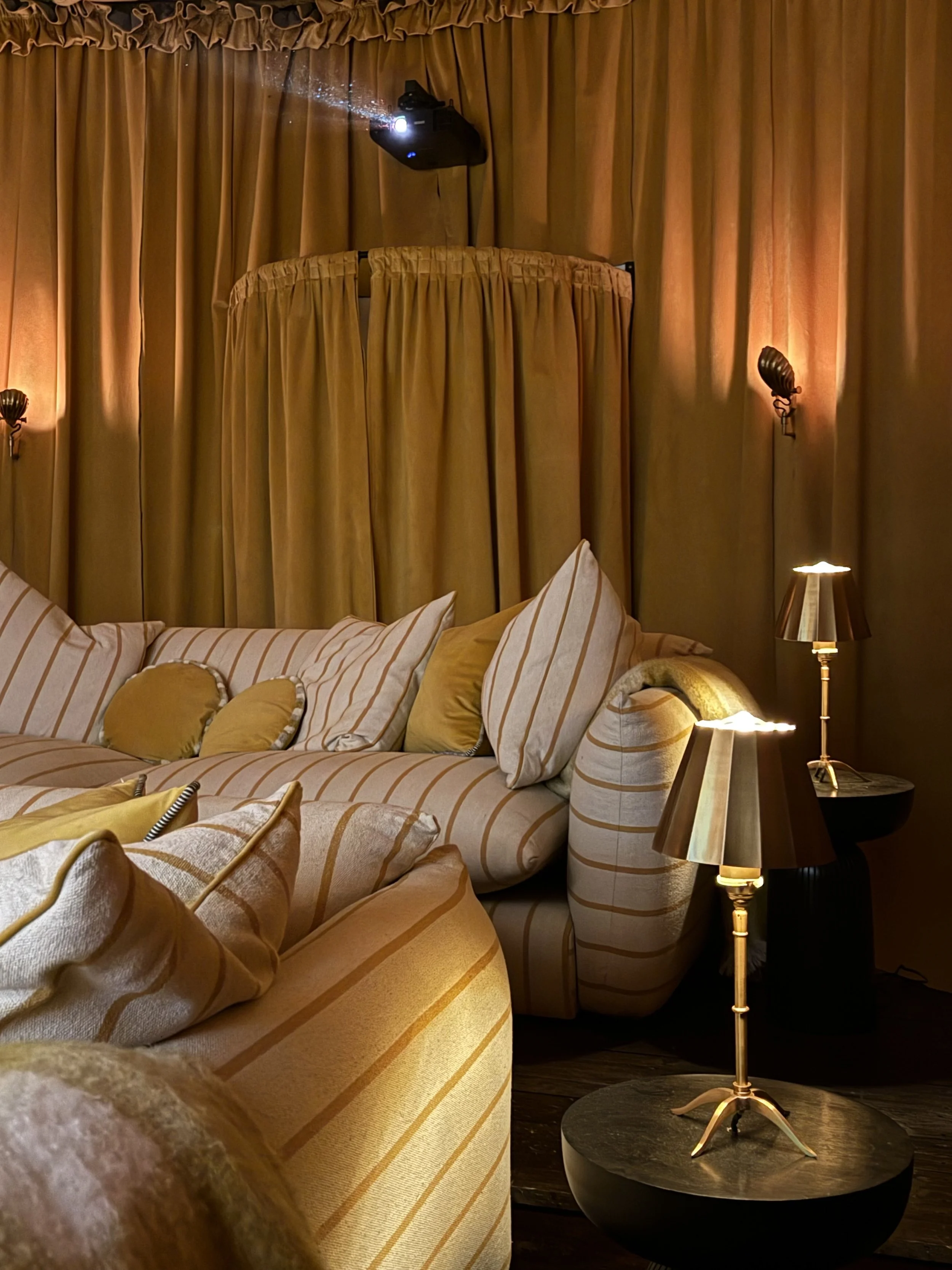 A cozy living room corner with a striped beige and white sofa, gold and beige pillows, a black side table with a gold lamp, and a wall-mounted lamp. The room has gold curtains and a projector mounted on the ceiling.