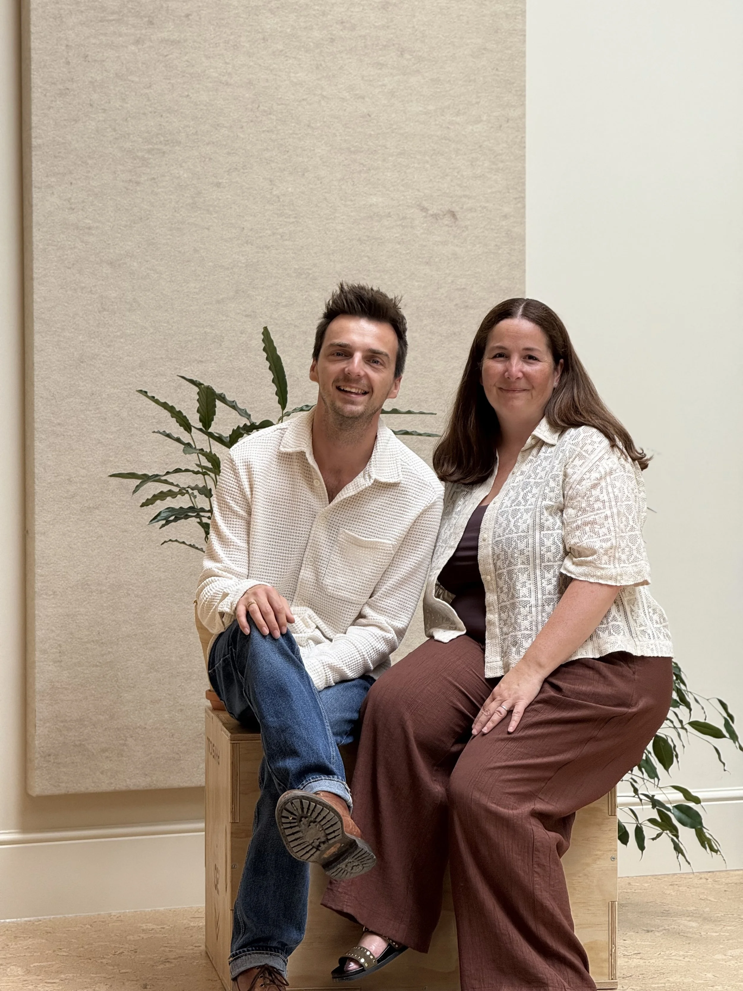 A man and woman sitting together on a wooden bench, smiling, with a plant and beige wall in the background.