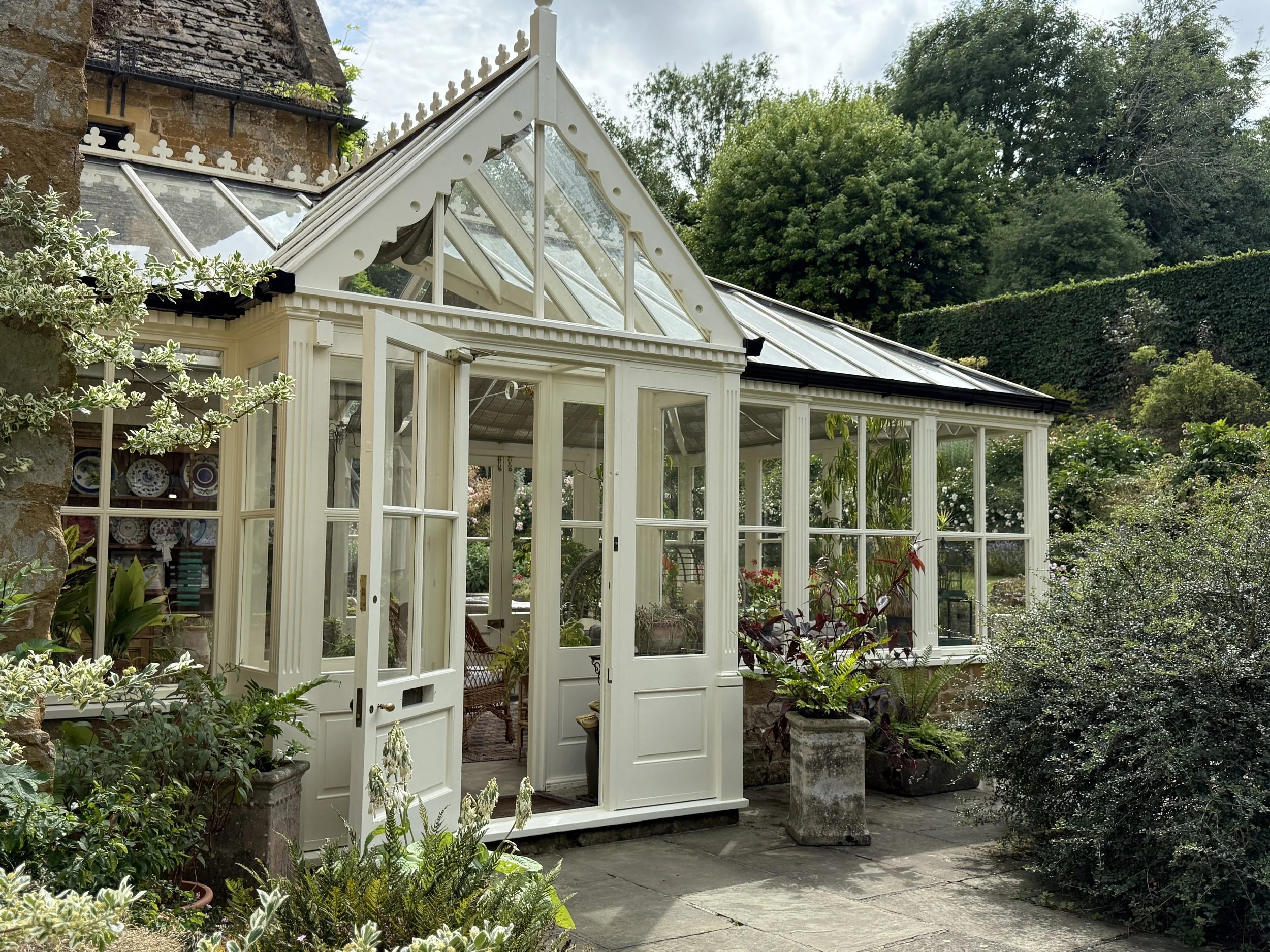 A traditional greenhouse with white-painted wooden framing and glass panels, situated in a lush garden with greenery and flowering plants.