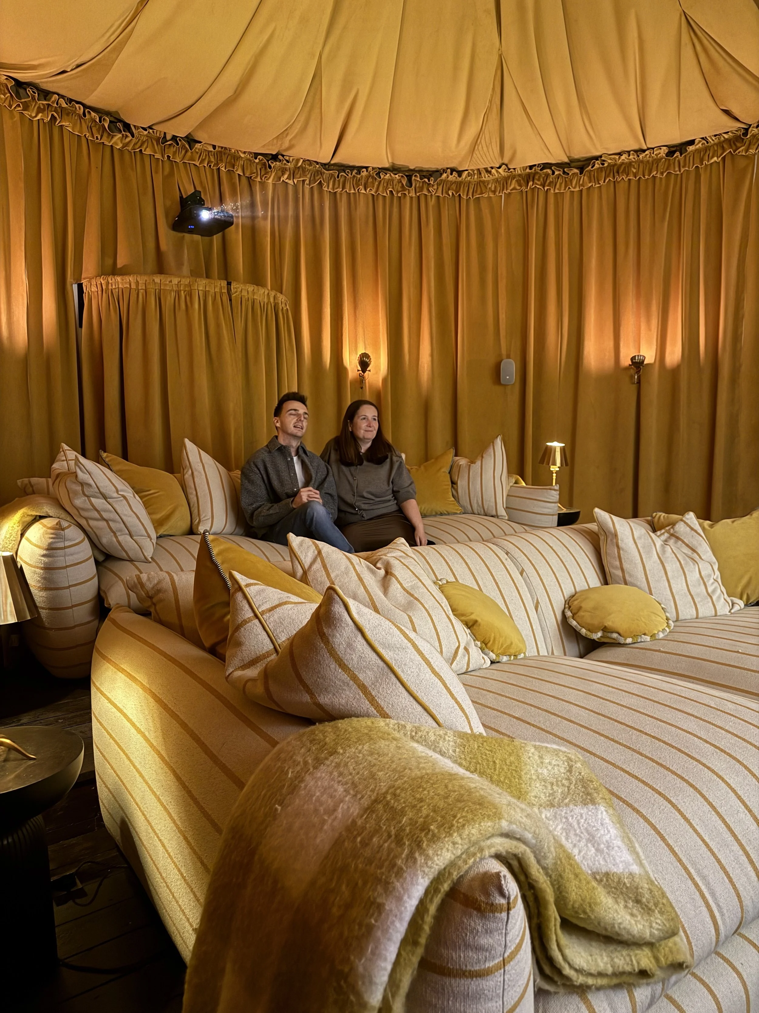 Two people sitting on a large, striped yellow and beige sectional sofa in a room with mustard-colored curtains and pillows, with dim warm lighting and a projector mounted on the ceiling.