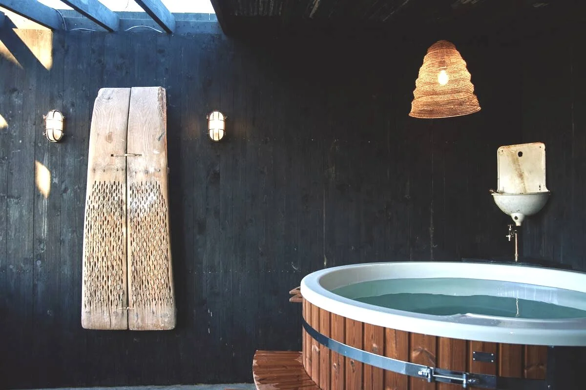 Indoor hot tub with wooden exterior, dark wooden wall, woven lamp, wall-mounted basin sink, and wall-mounted lights.