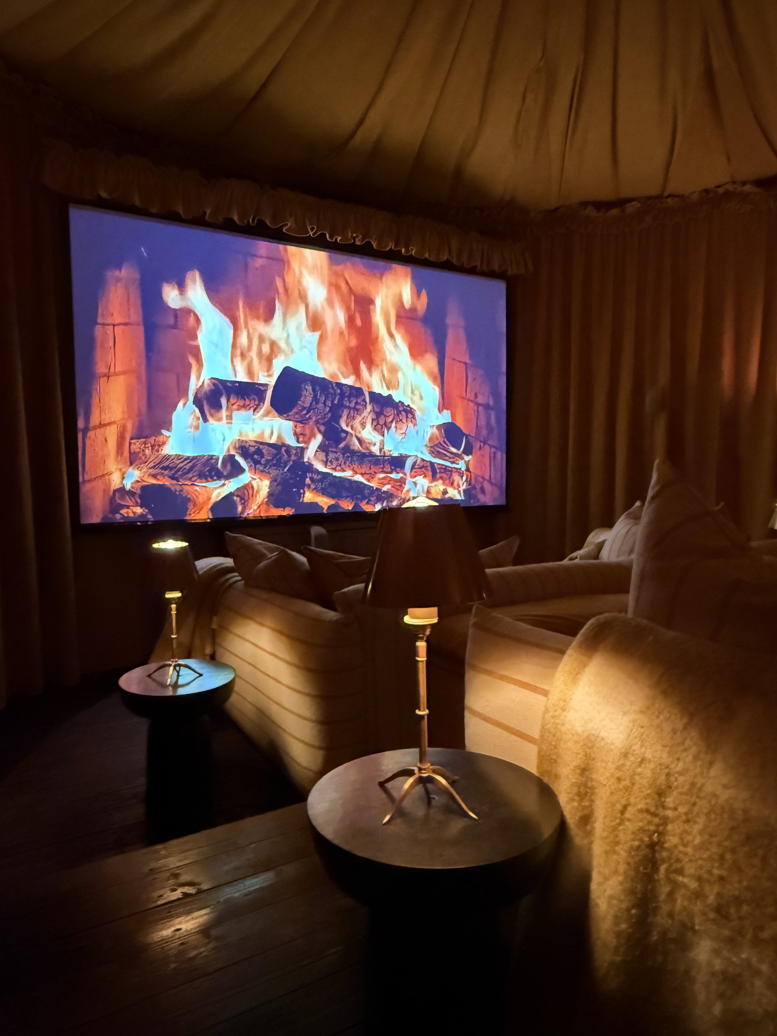 Living room with a large fireplace display on a screen, showing animated flames and logs. The room has curtains, sofas, and small tables with lamps.