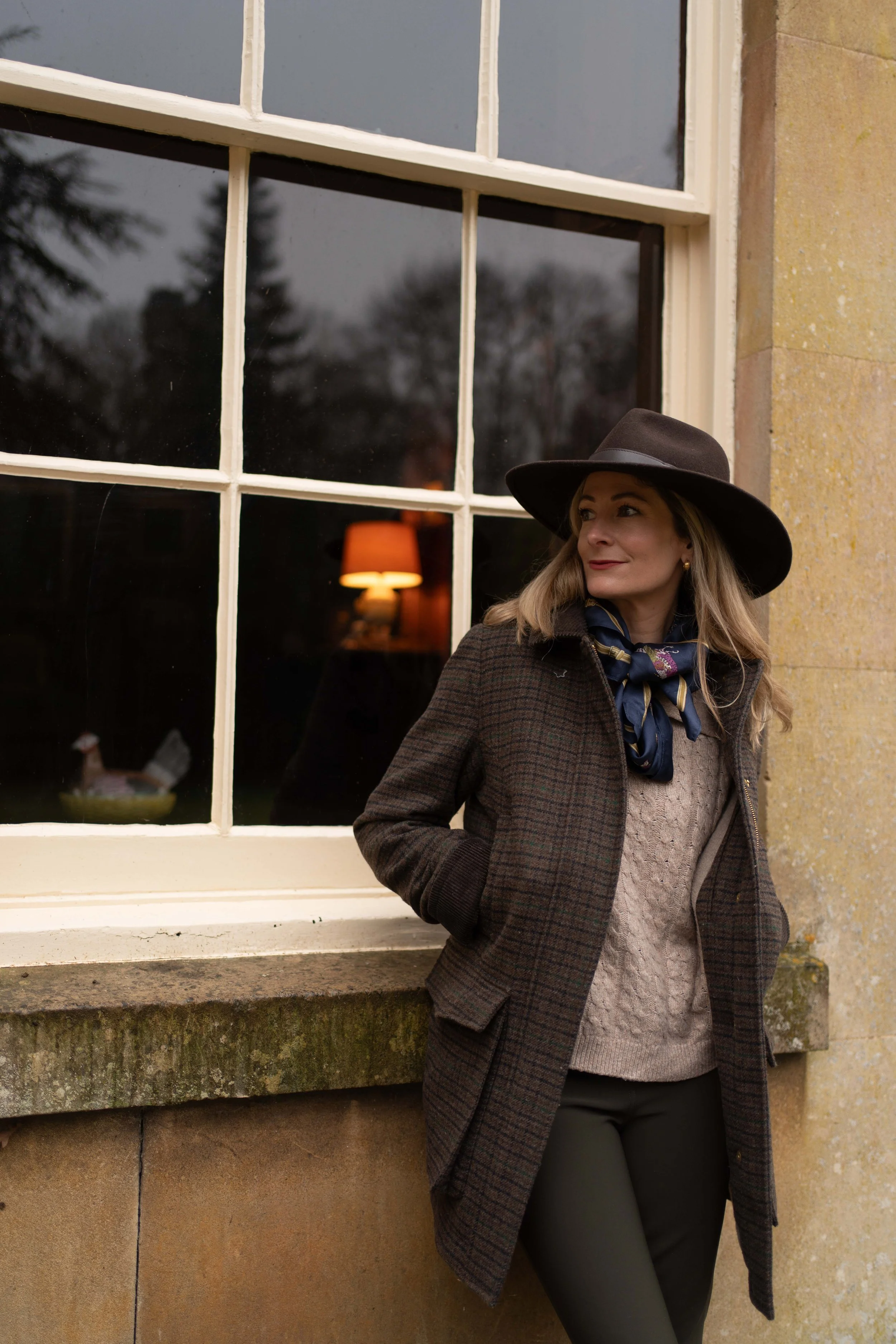 A woman outdoors leaning against a stone wall beside a window, wearing a wide-brimmed hat, a plaid coat, a sweater, and a scarf.