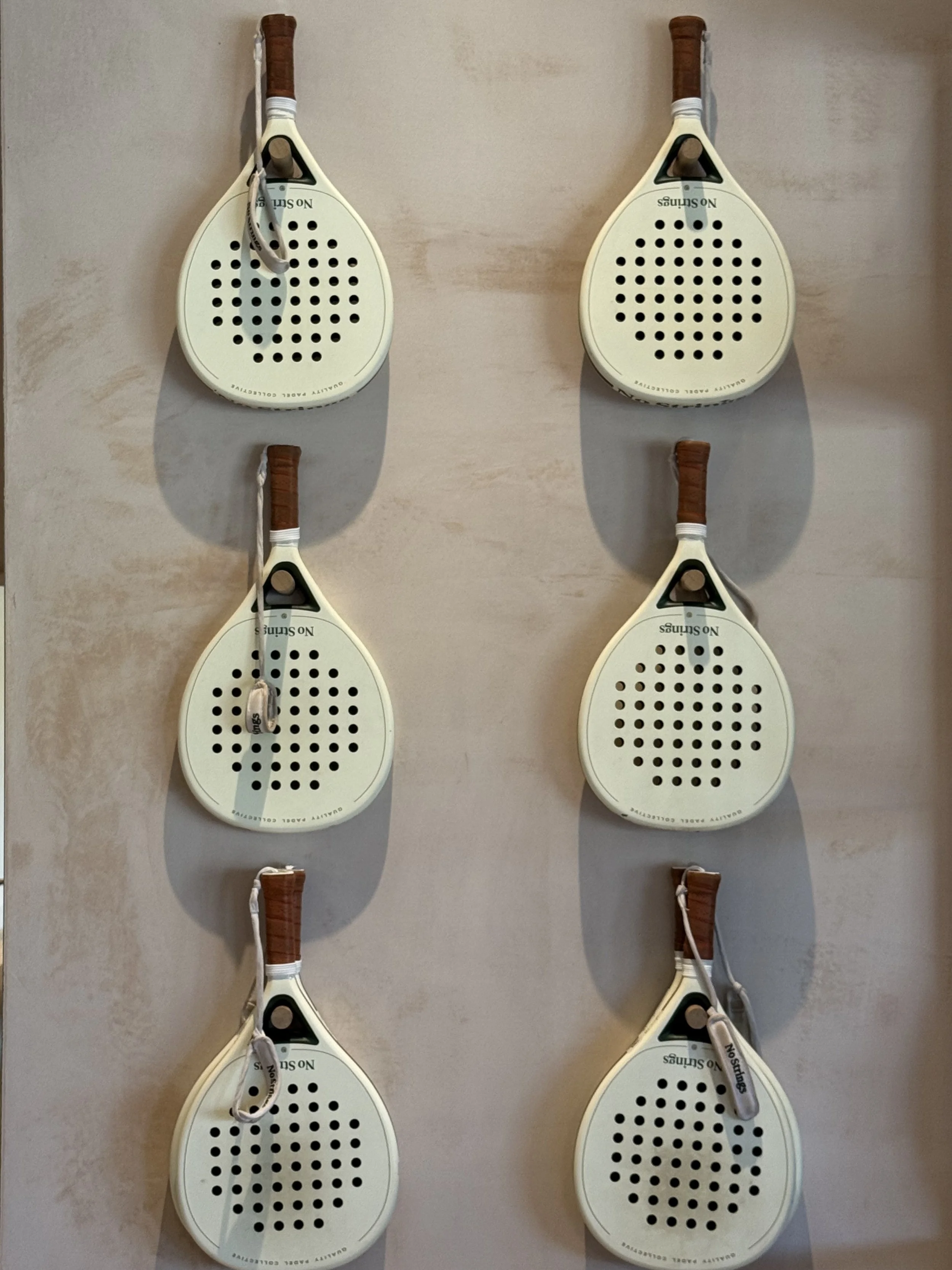 Six vintage white paddle tennis rackets mounted on a wall in two columns of three, each with brown grips and a grey wrist strap.