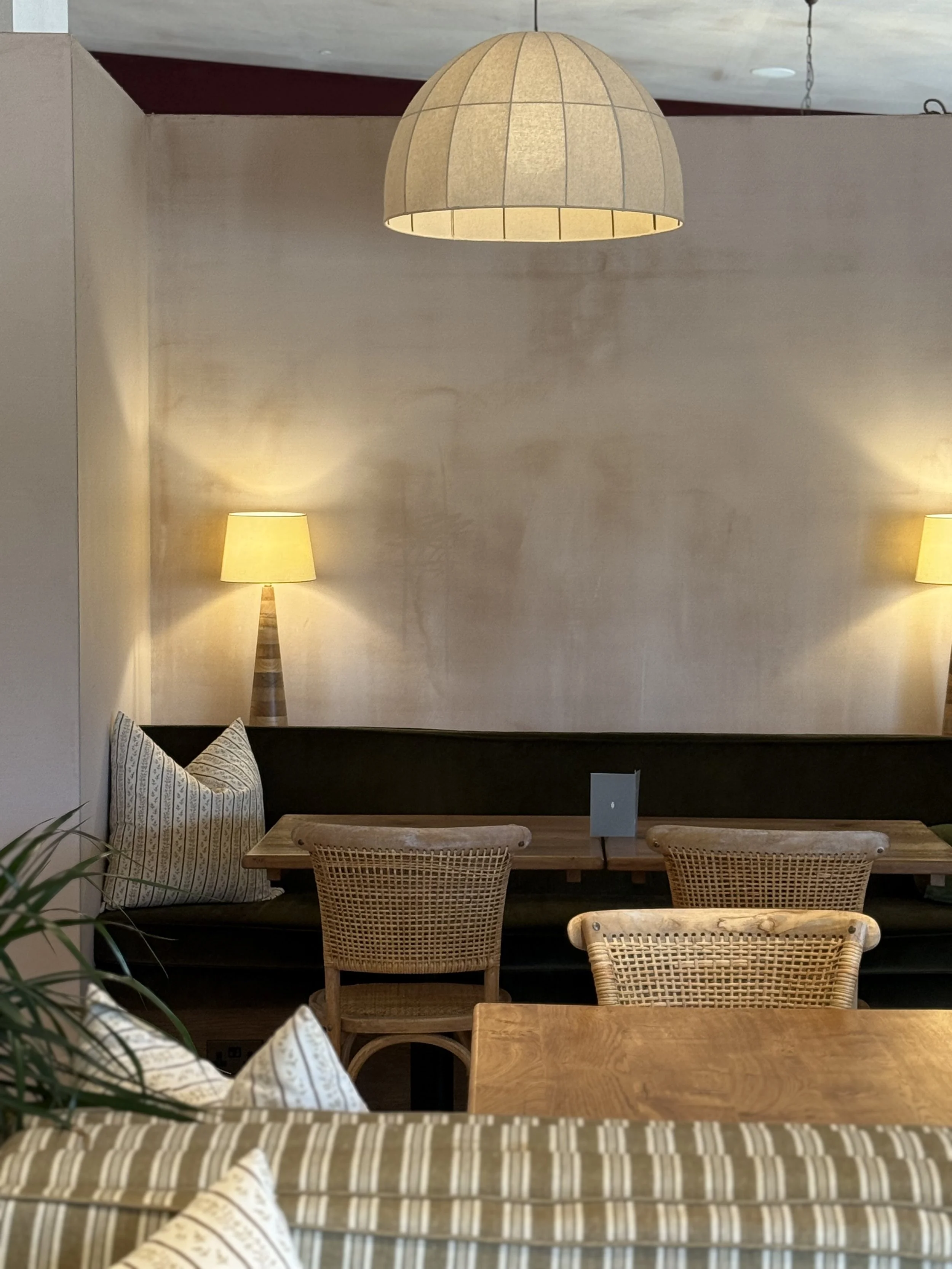Interior of a cozy cafe with wooden tables and woven chairs, a black cushioned bench with striped pillows, two wall-mounted lamps, and a hanging pendant light on a beige wall.
