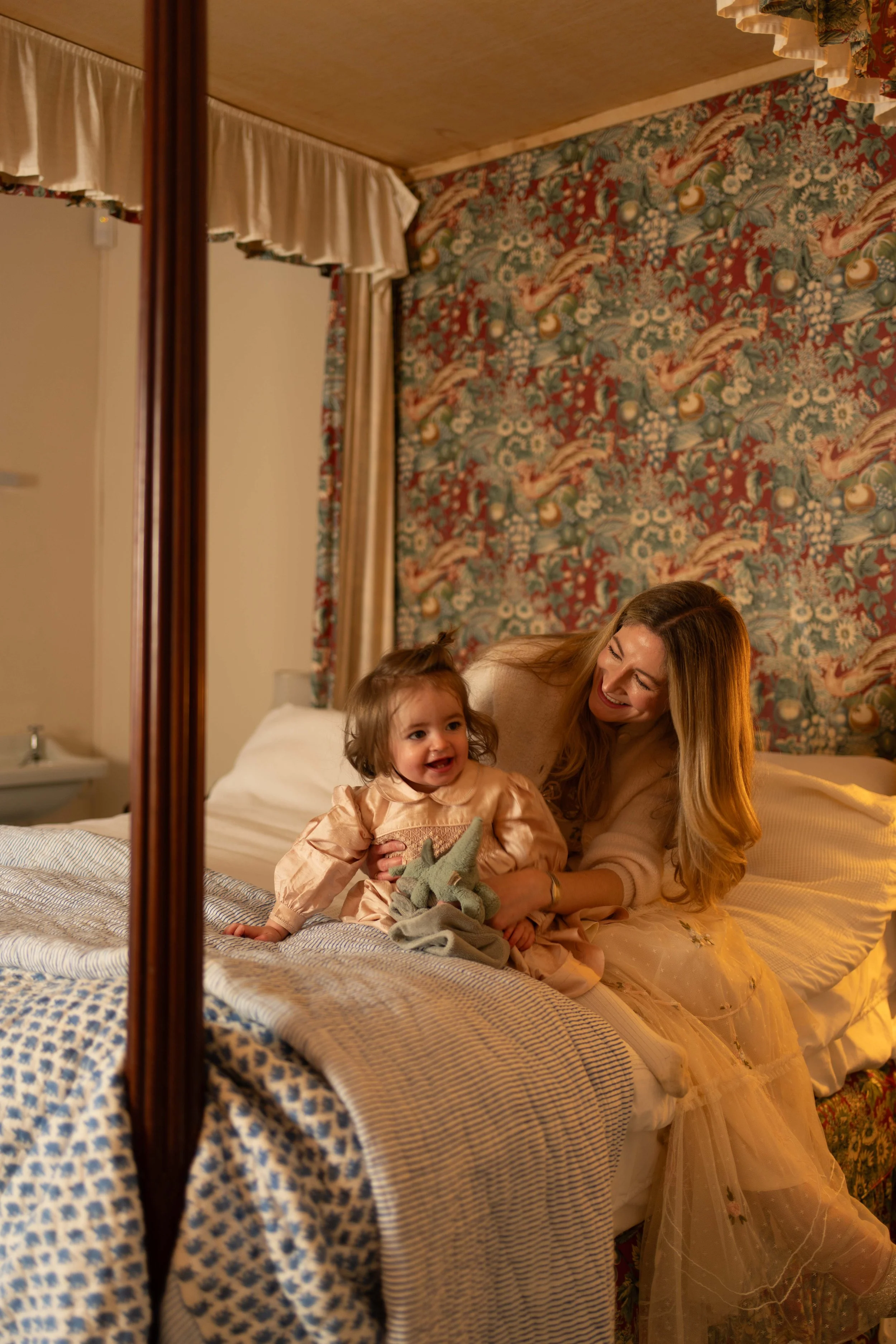 A woman and a young girl sitting on a bed, smiling and laughing together in a bedroom with a vintage floral wallpaper and curtains.