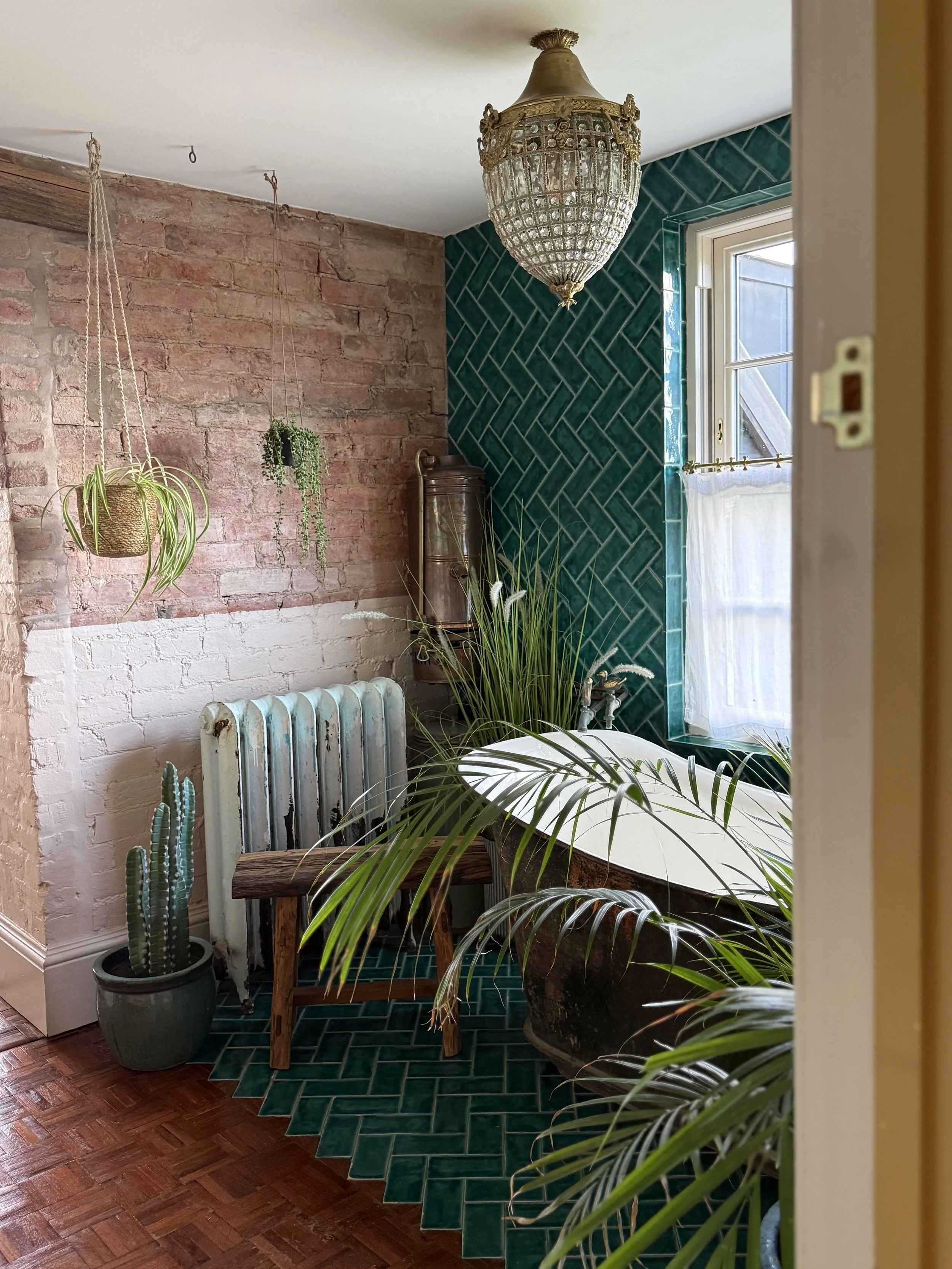 A vintage bathtub with greenery in the bathroom, featuring a brick wall, teal tiled wall, hanging plants, an old radiator, and a window with white curtains.