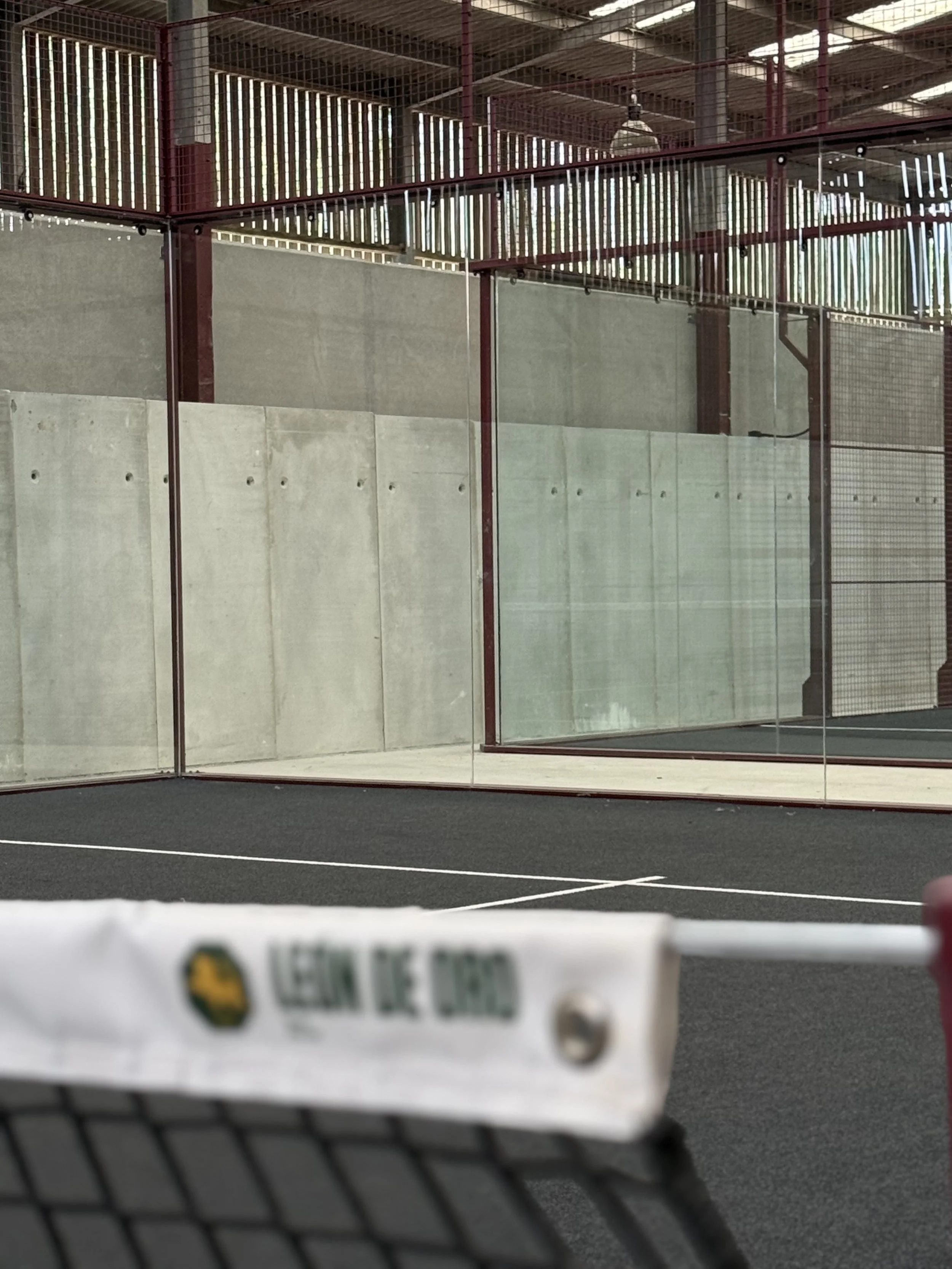 Indoor sports court with glass walls, red framing, and a black surface. Parts of a tennis racket and ball are visible in the foreground.