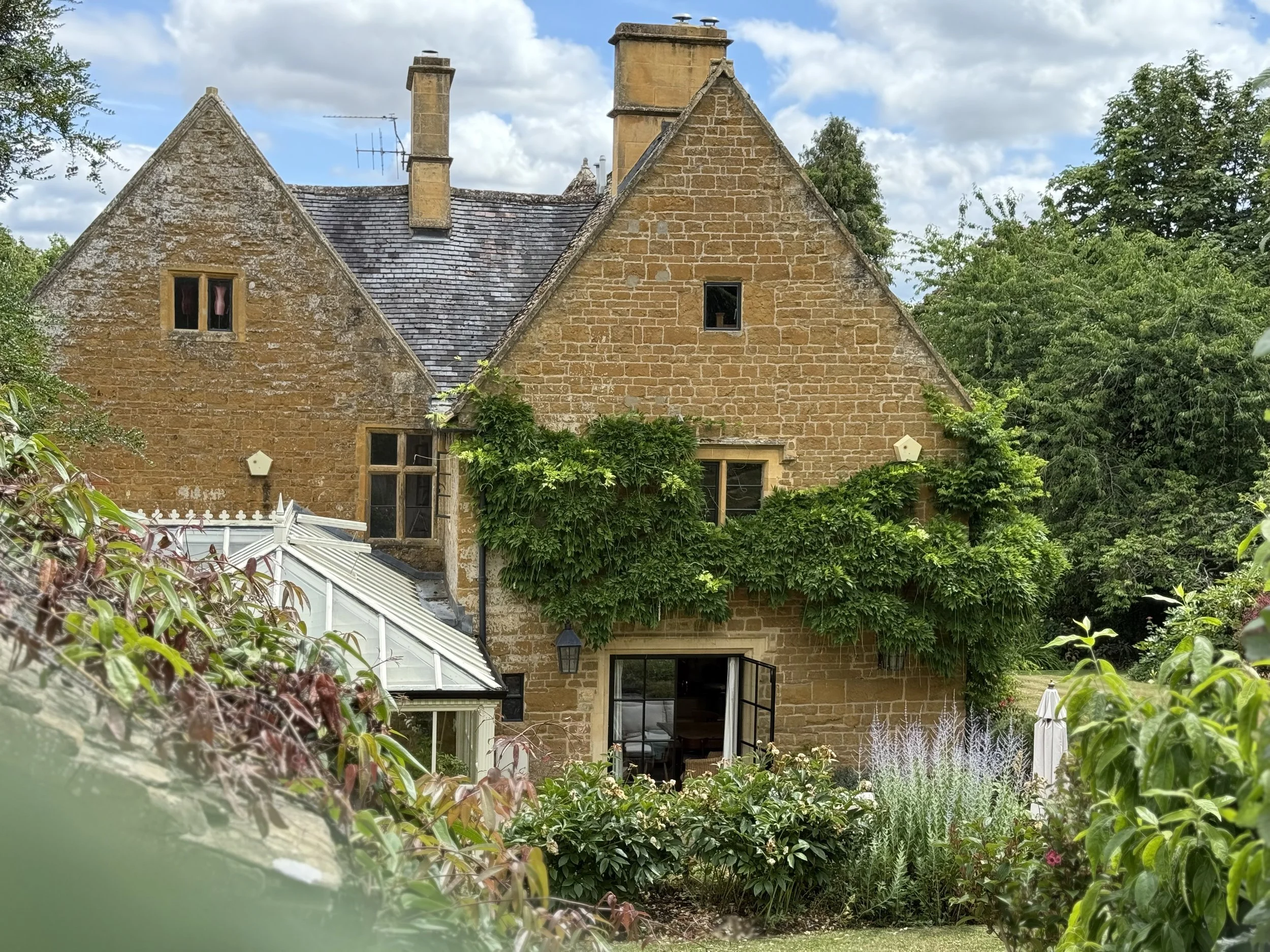 Old stone house with greenery and plants growing on its facade, windows, and a glass conservatory attached, surrounded by trees and plants, under a partly cloudy sky.