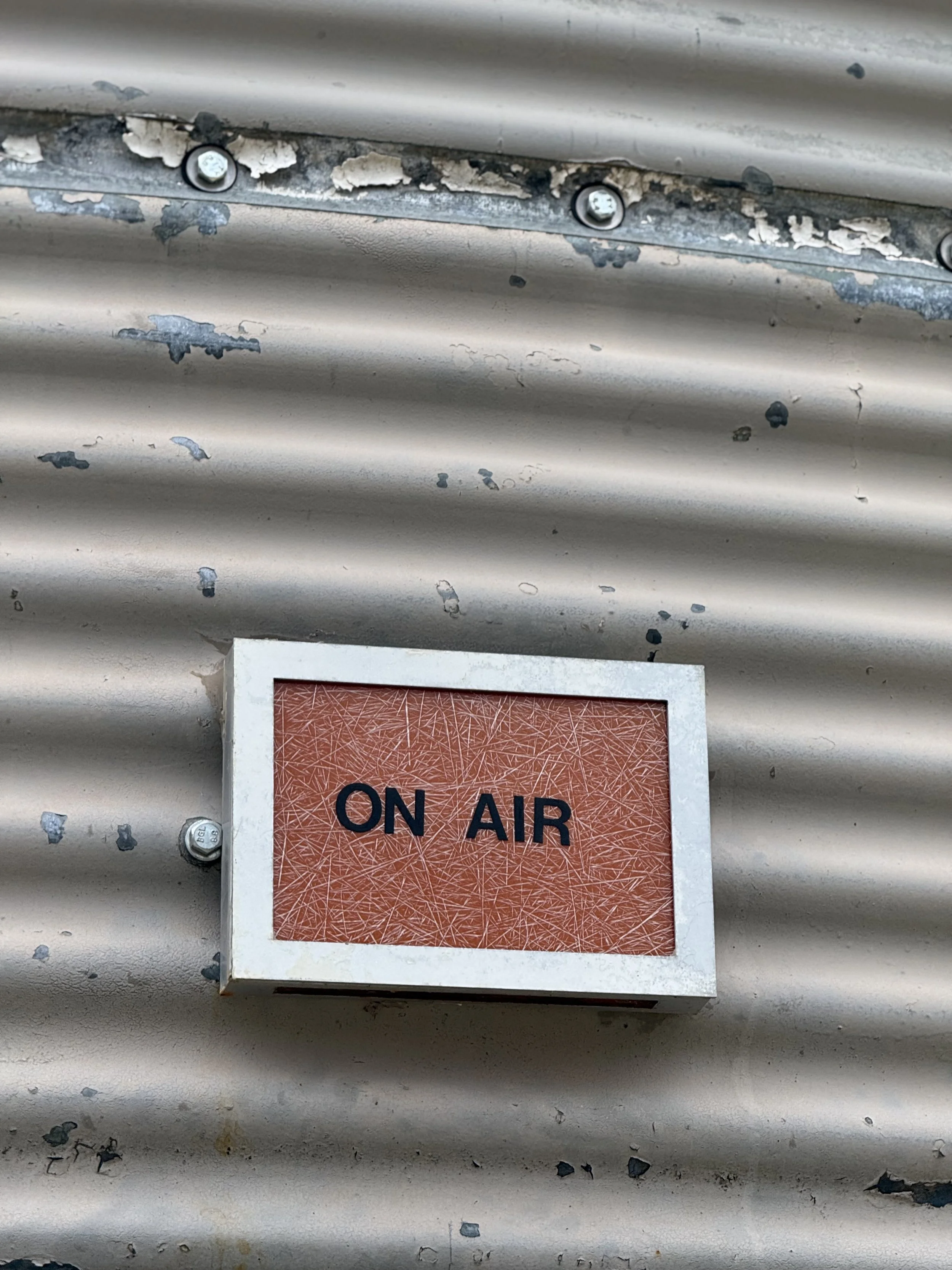 Close-up of a sign that reads 'ON AIR' on a metallic corrugated wall with peeling paint and screws.