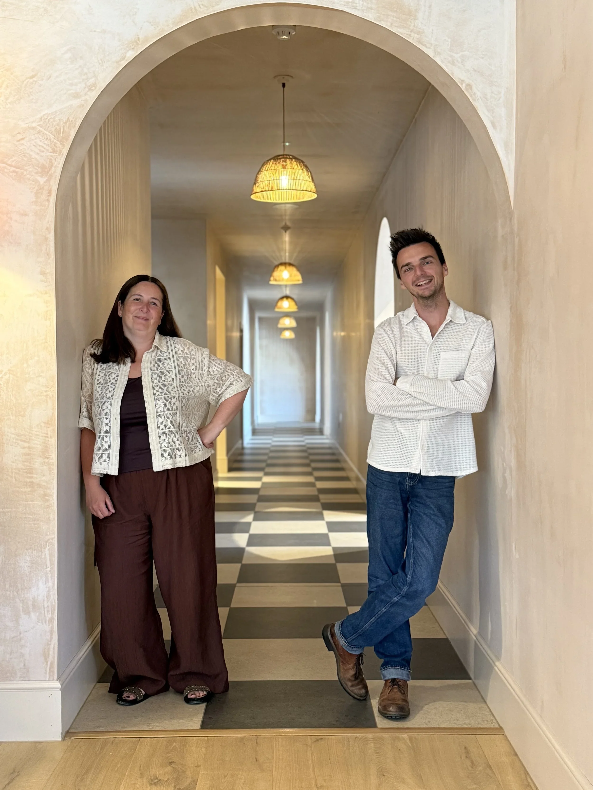 A woman and a man standing in a hallway with arched openings, wall-mounted lamps, and checkered flooring. The woman is on the left, wearing a patterned cardigan, dark shirt, and loose brown pants with sandals. The man is on the right, wearing a white checkered shirt, blue jeans, and brown boots, leaning against the wall with arms crossed and smiling.