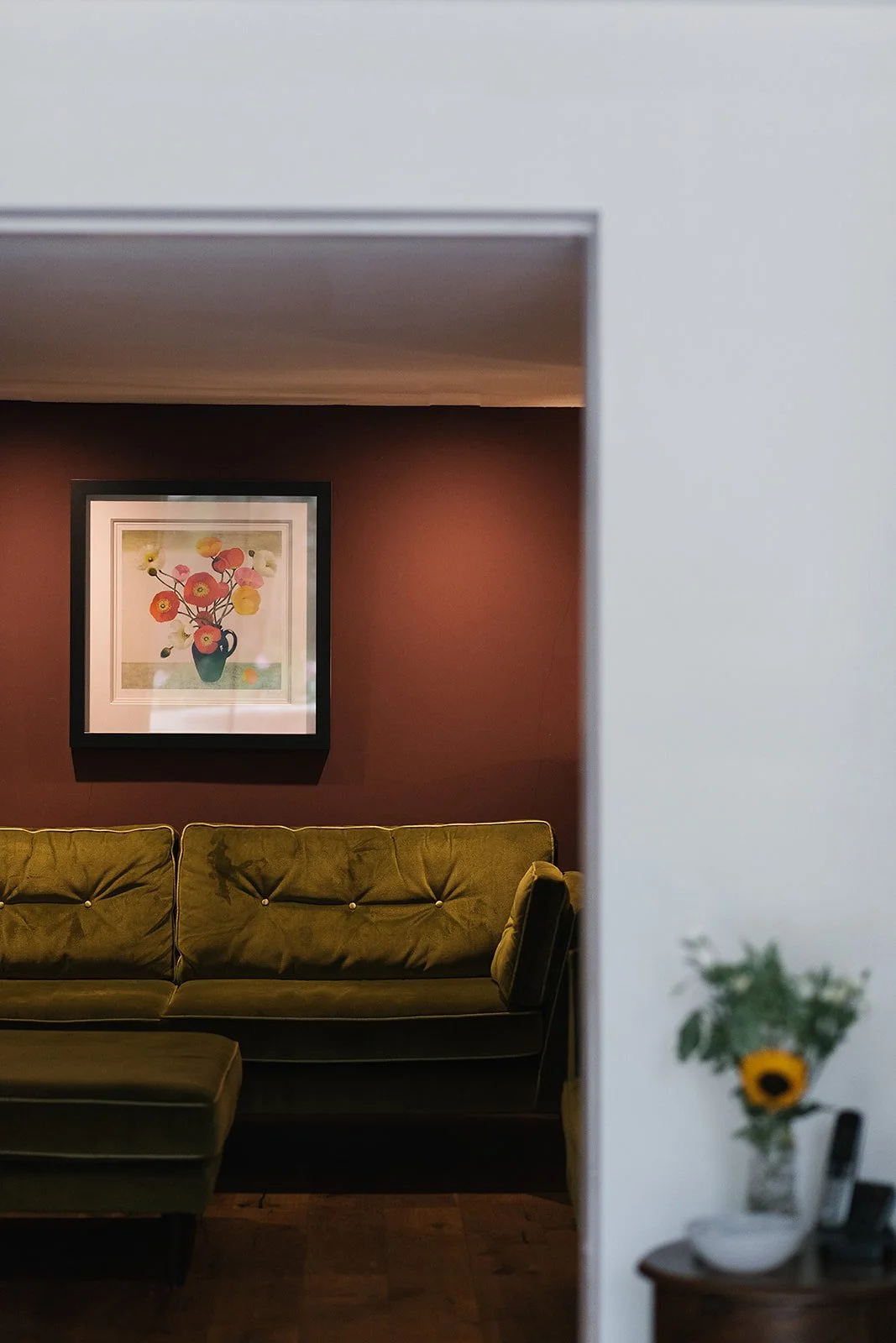 A view of a cozy living room through a partial doorway, featuring a green velvet sofa, a framed floral painting on a dark brown wall, and a side table with a sunflower bouquet in a vase.