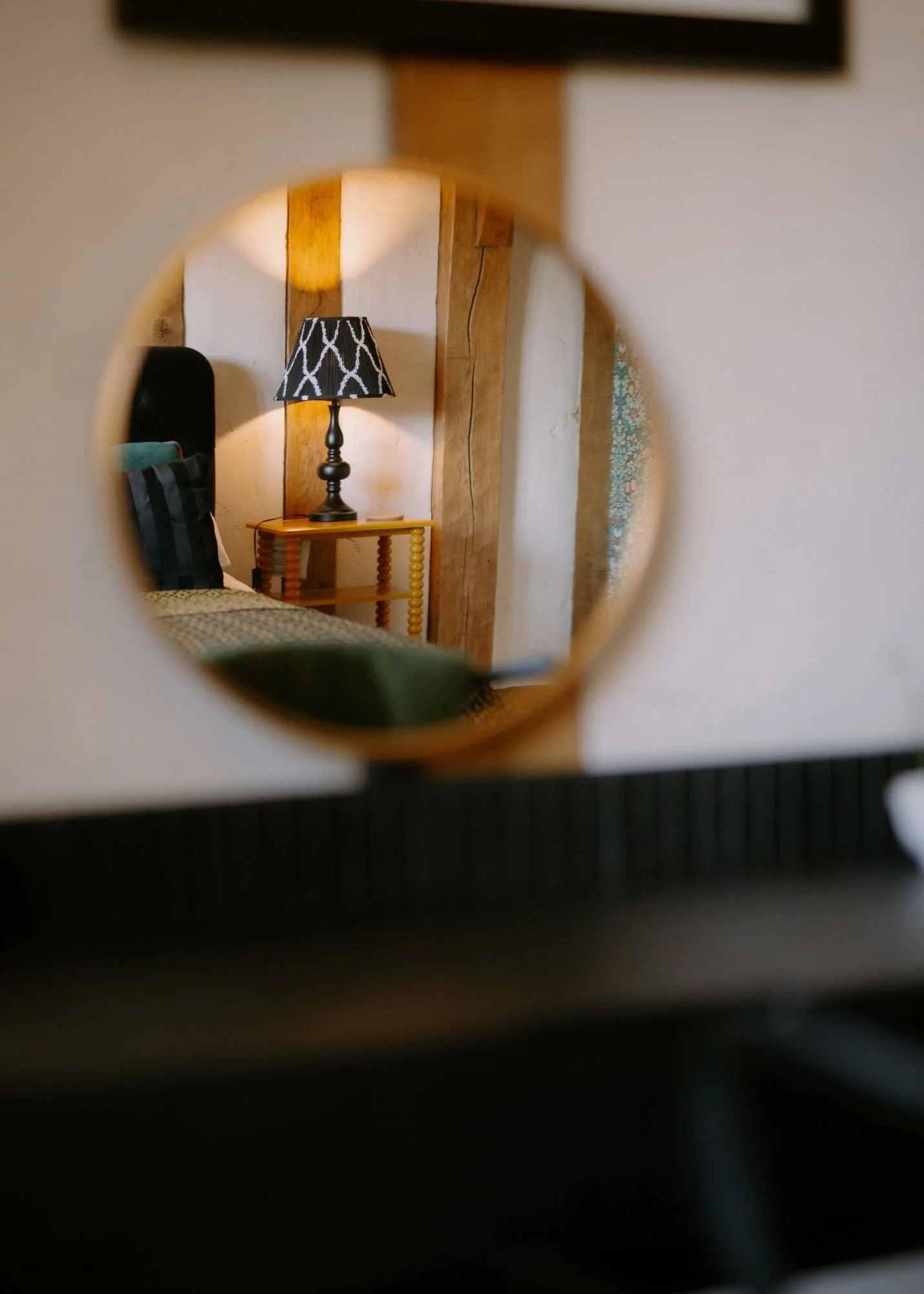A bedroom view through a round mirror, showing a black and white patterned lampshade on a bedside table and parts of a bed with black pillows, wood beams, and a textured wall.