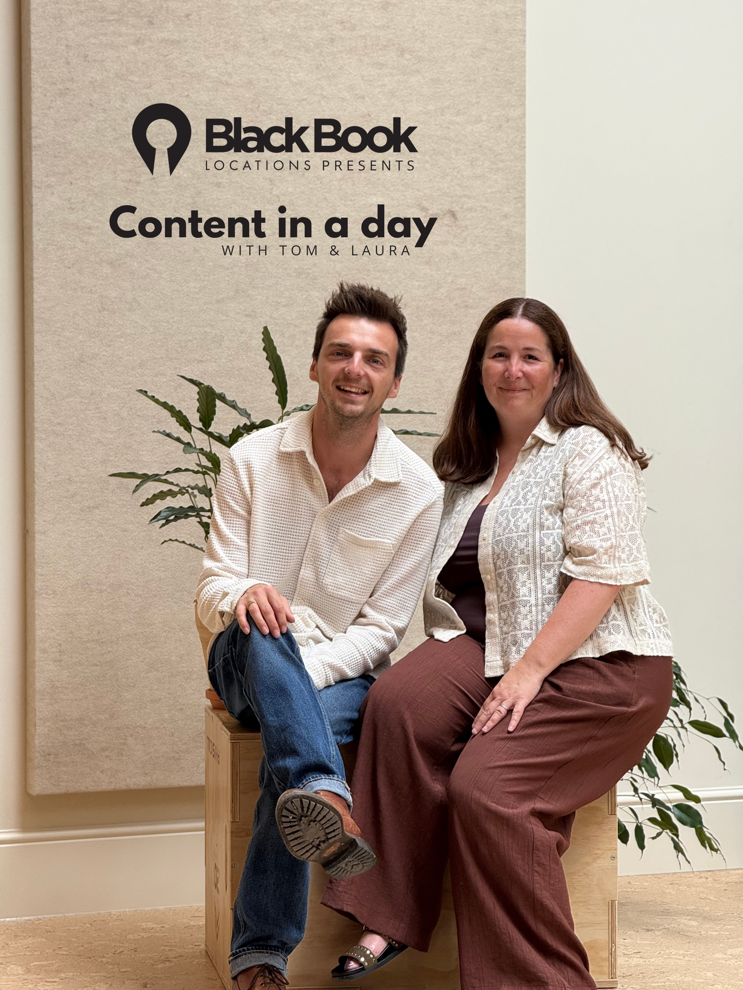 A man and woman sitting and smiling in front of a beige wall with a black logo and text. There is a green plant behind the man.