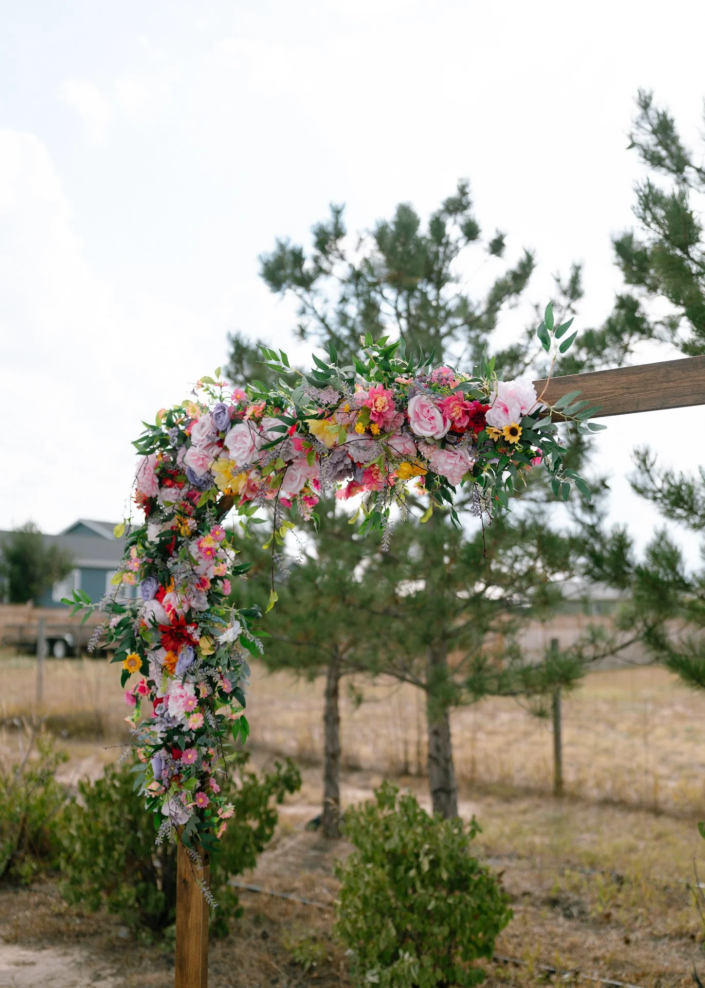 Floral arch with pink and purple flowers and green leaves outdoors, with trees and houses in the background.