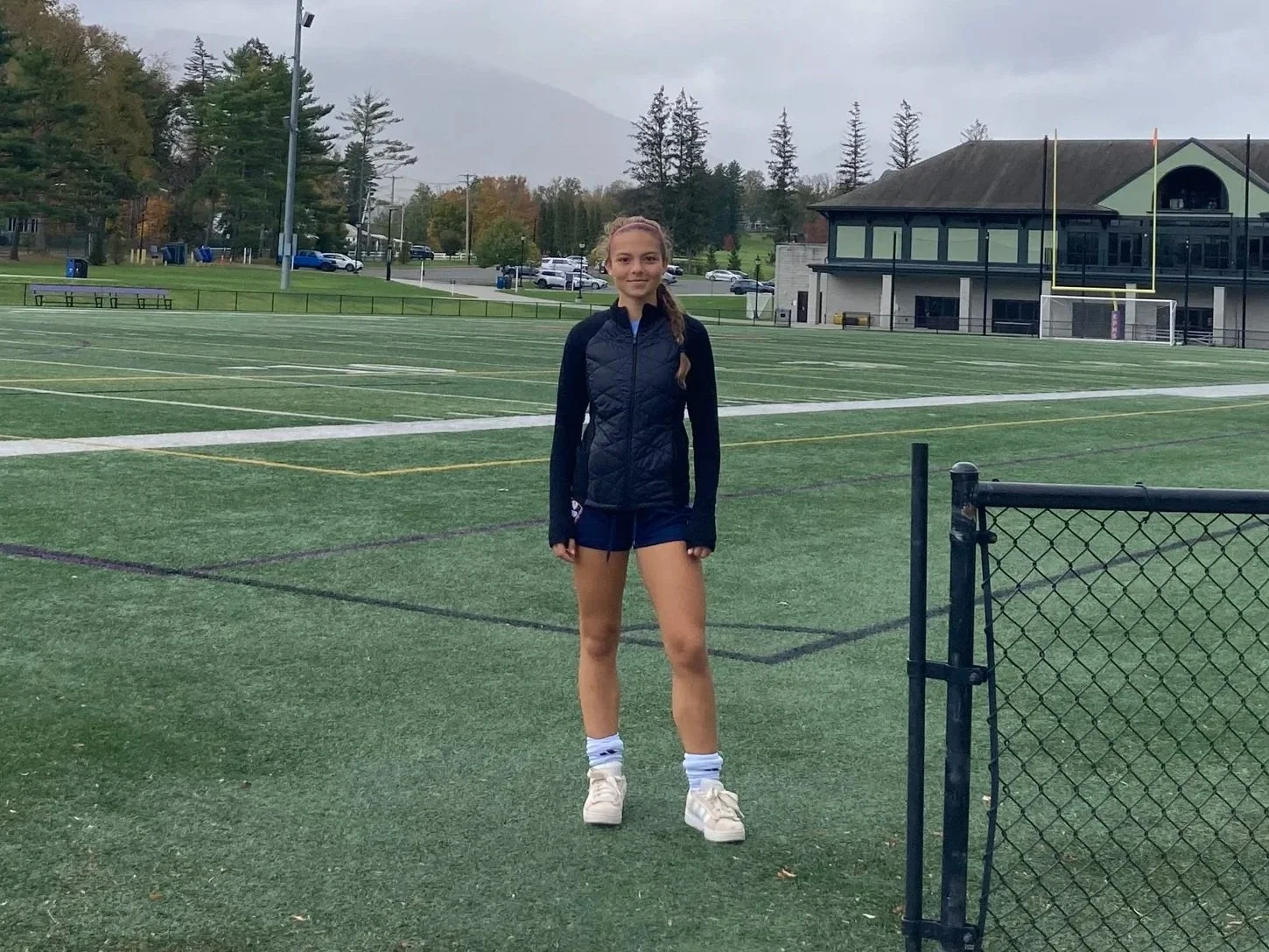 Natalija standing on a soccer field on a cloudy day with trees, cars, a building, and a mountain in the background.