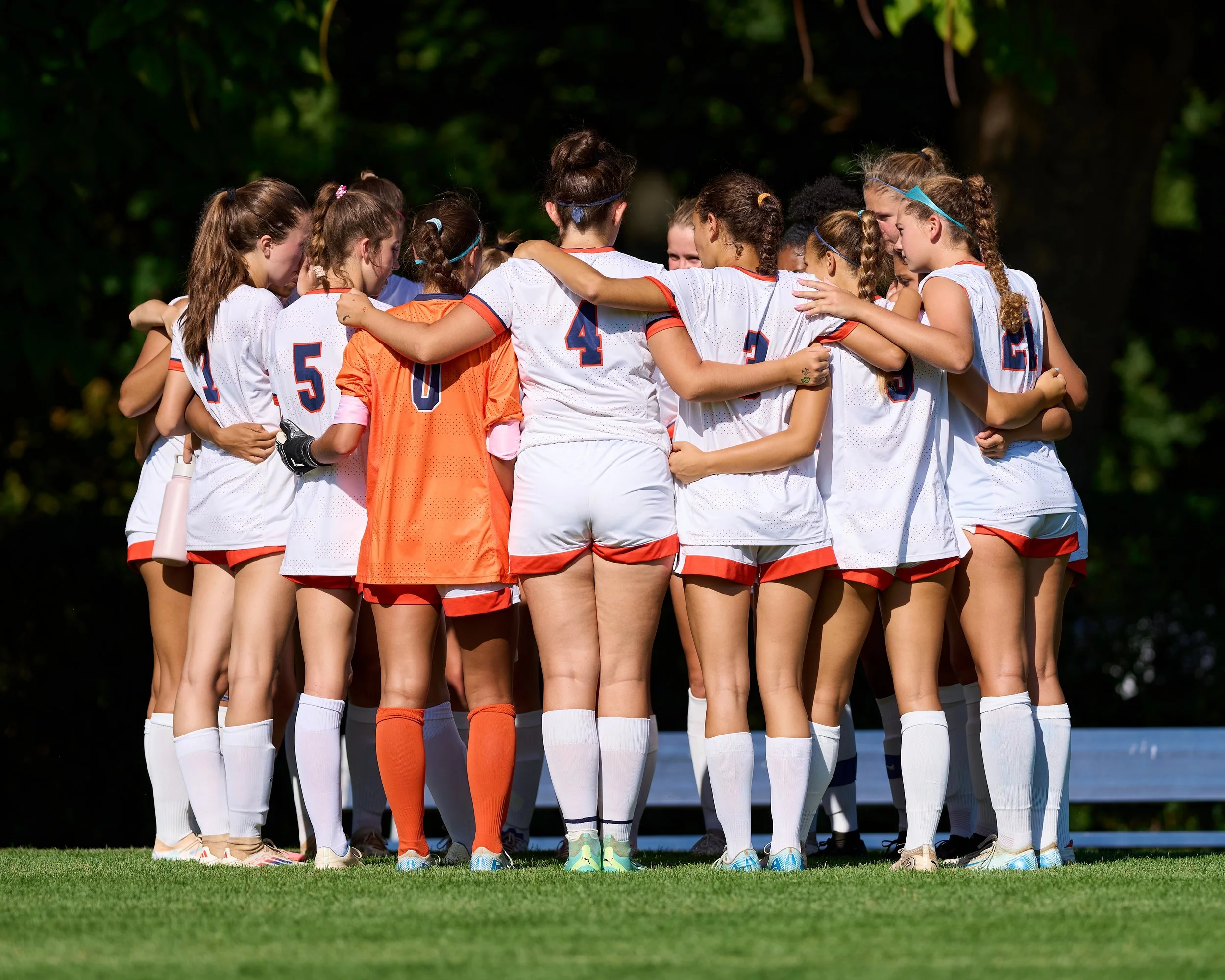 A group of female soccer players in white and orange uniforms huddled together on a soccer field, embracing each other in a team cheer.