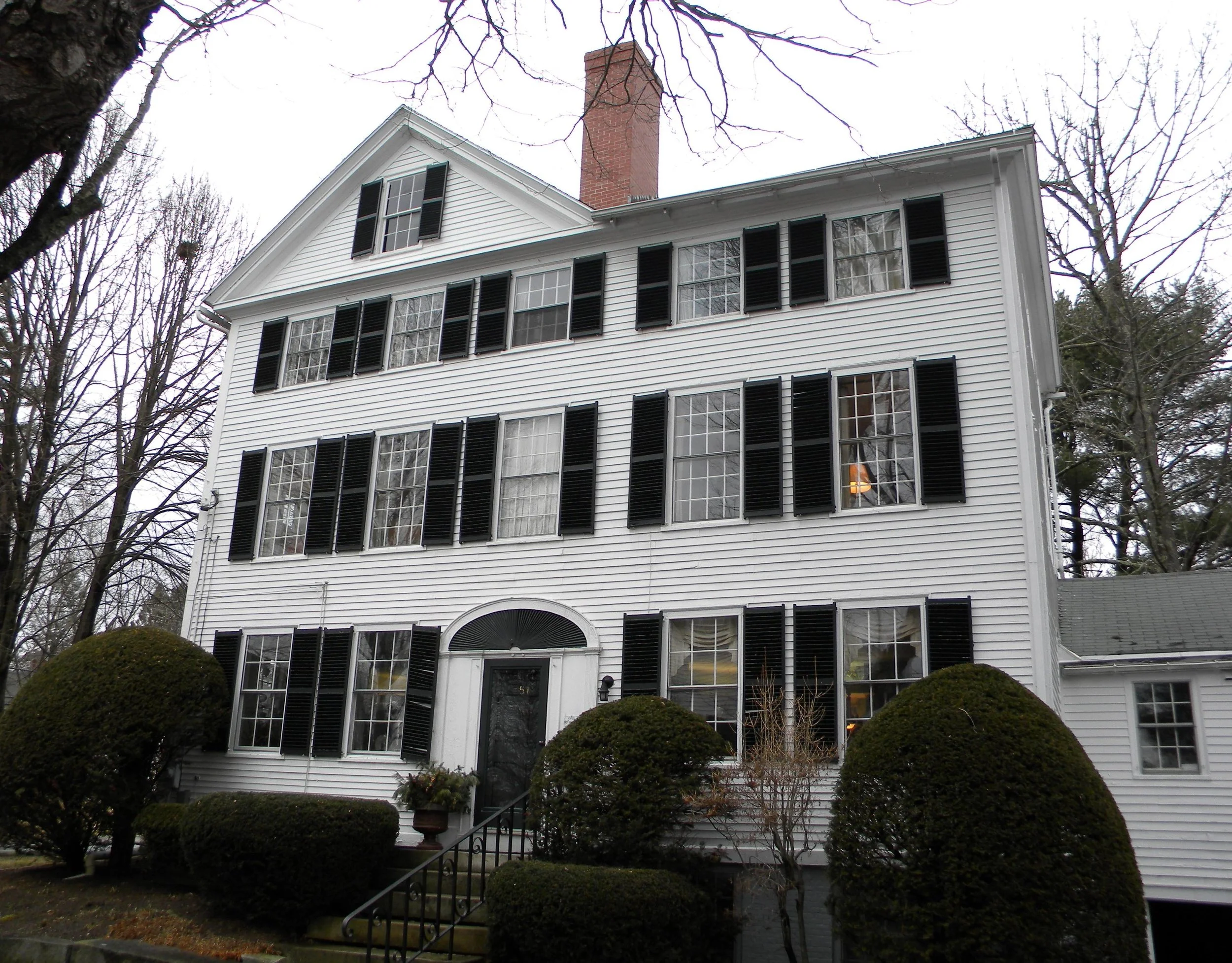 White multi-story house with black shutters and front steps, surrounded by bushes.