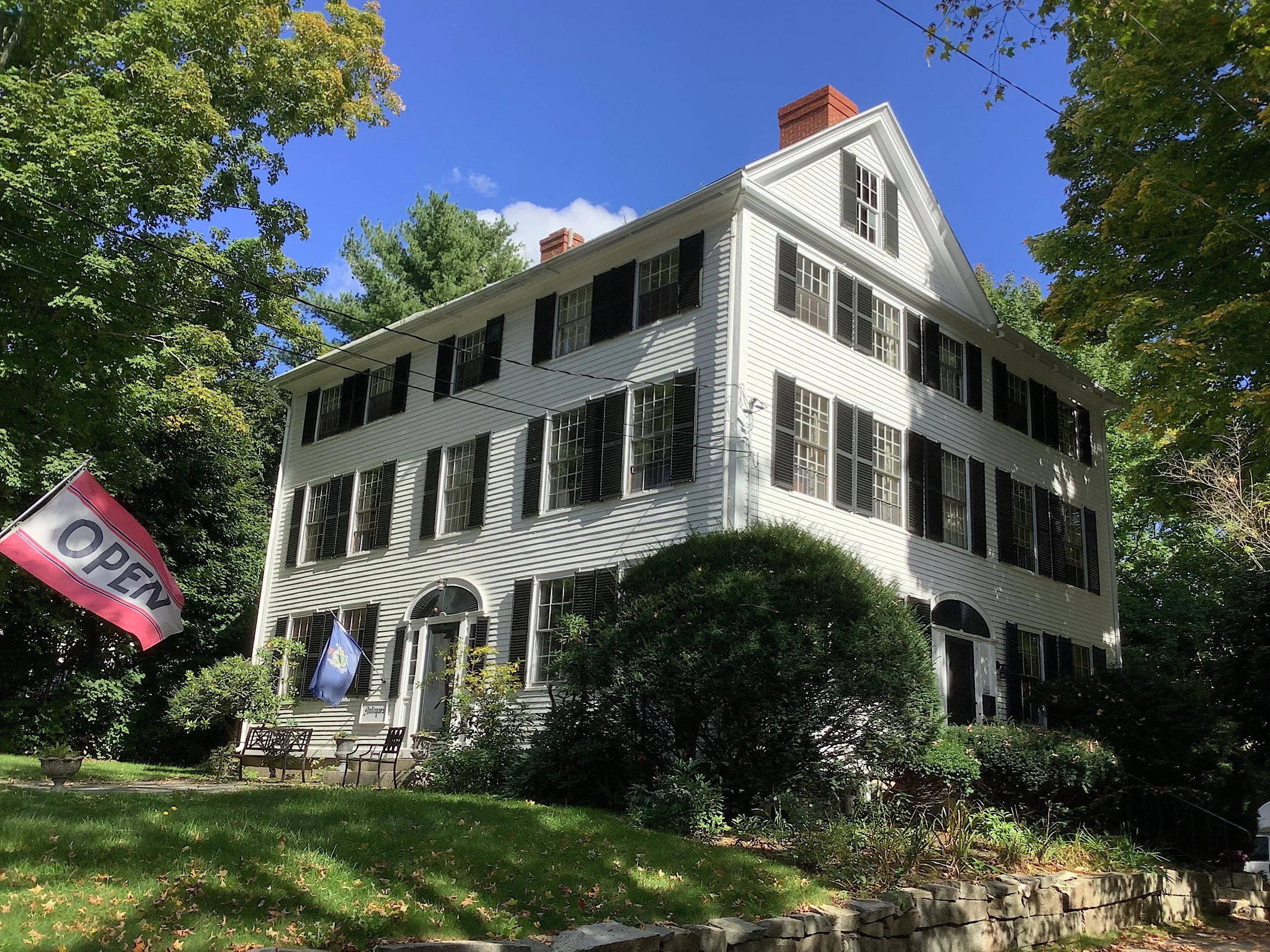 A large three-story white house with black shutters, surrounded by greenery and trees, with a brick chimney, a front porch with outdoor furniture, and a flag that says 'Open'.