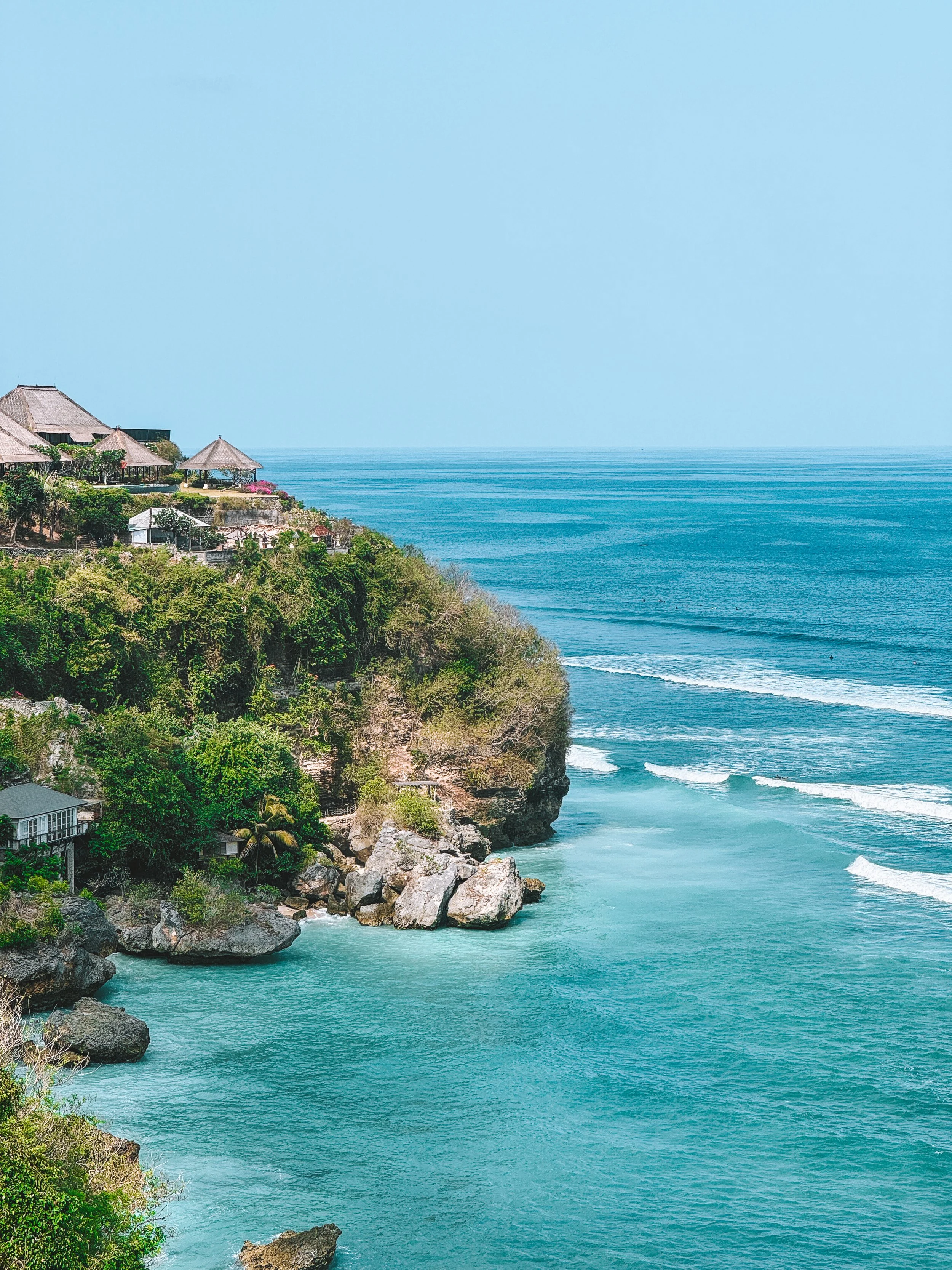 A coastal view with turquoise water, rocky cliffs, green vegetation, and buildings with thatched roofs on the hillside, under a clear blue sky.