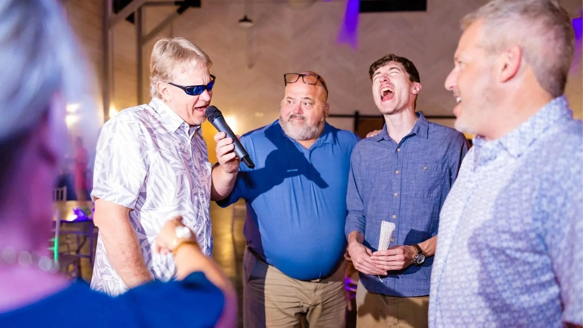 A group of middle-aged men are singing karaoke at a party, with one man holding a microphone and wearing sunglasses, and others smiling and laughing.