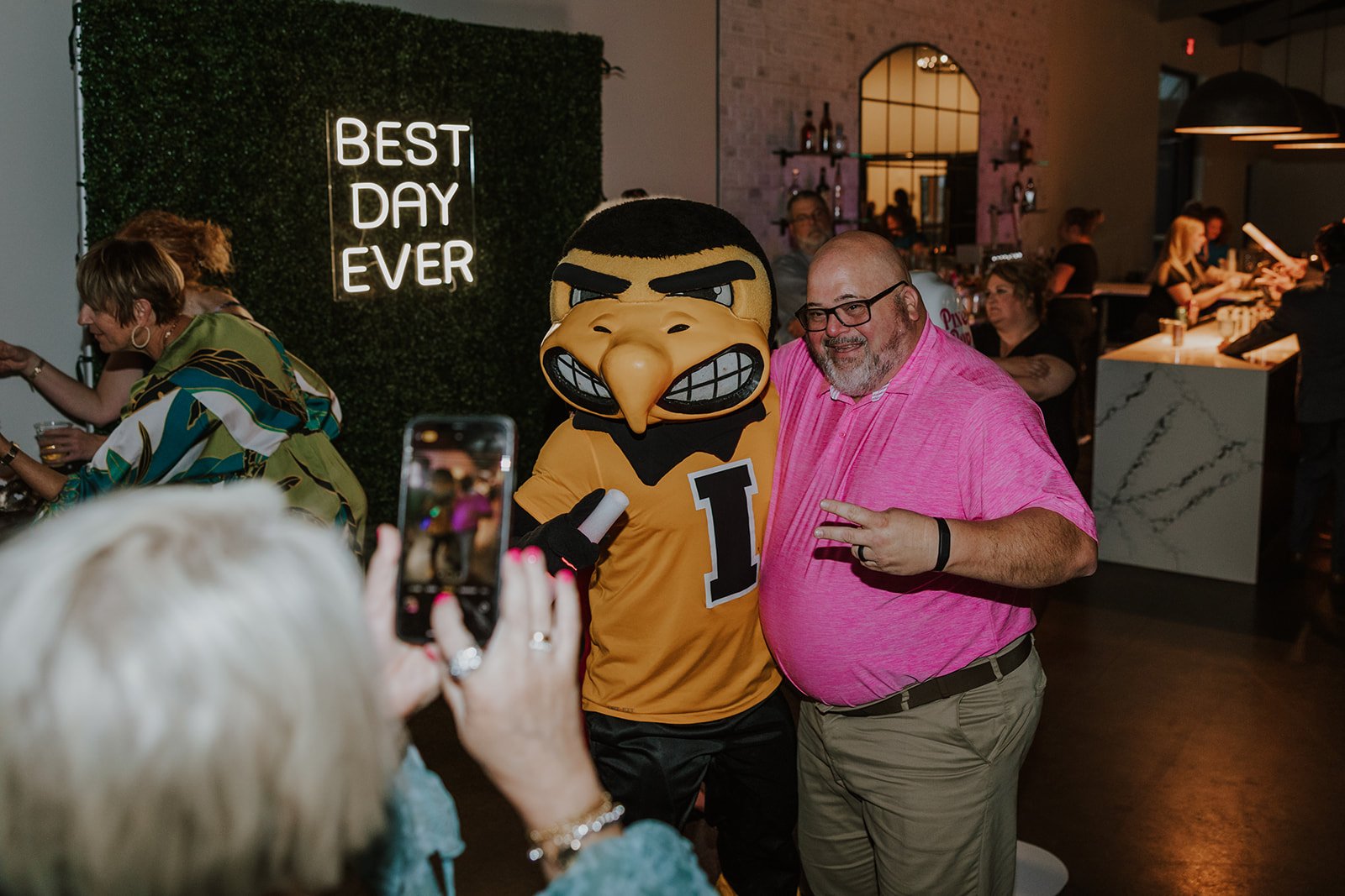 Person in a Florida Gators mascot costume posing with a man in a pink shirt at an indoor event. People are taking photos and socializing, with a neon sign that reads 'Best Day Ever' in the background.