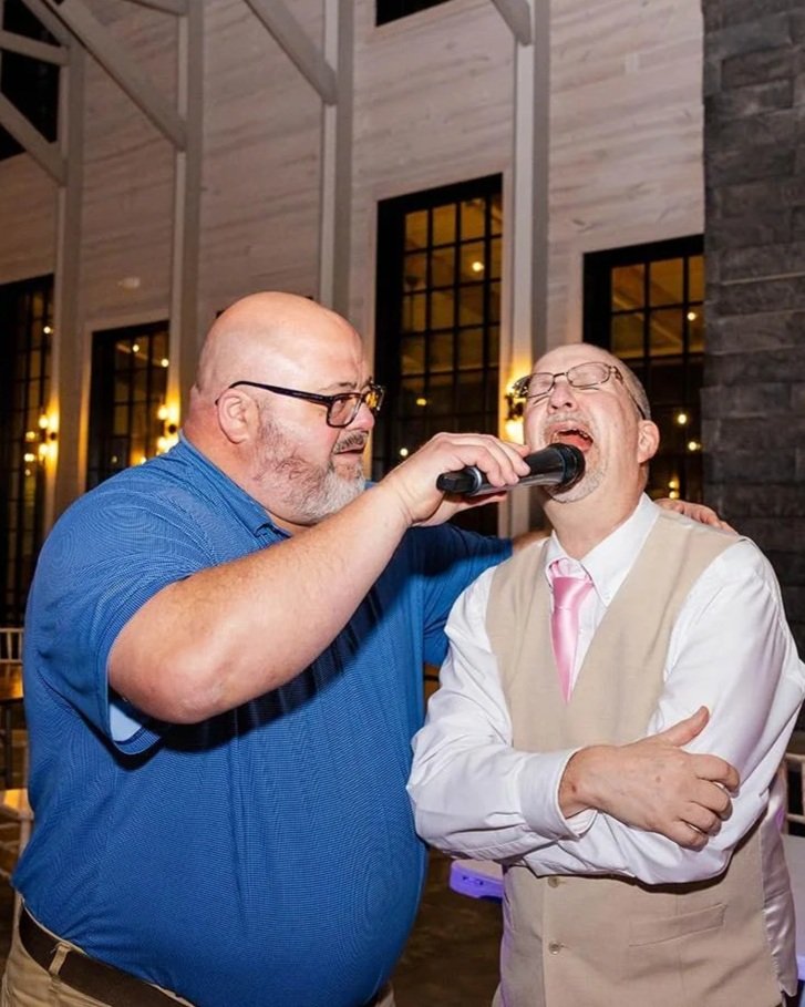 Two men dressed in formal attire, one in a blue shirt and the other in a beige vest with a pink tie, singing into a microphone at an indoor event with large windows and warm lighting.