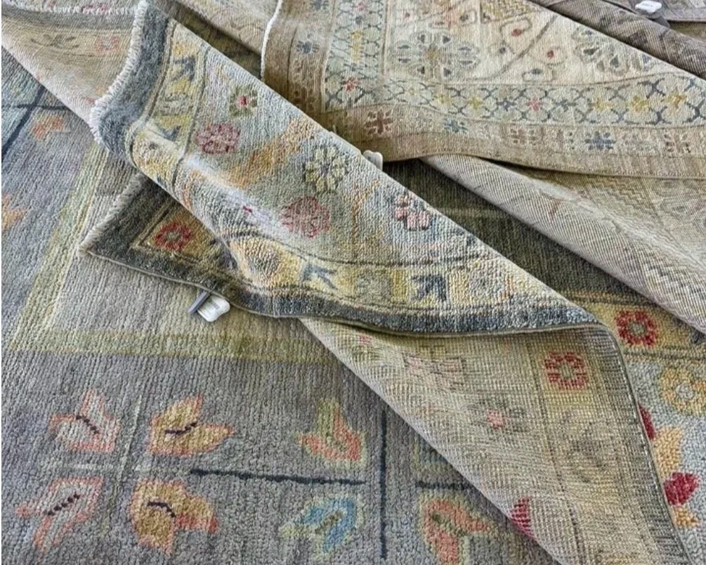 Close-up of a folded, patterned area rug with intricate designs and muted colors, partly revealing the underside of the rug.