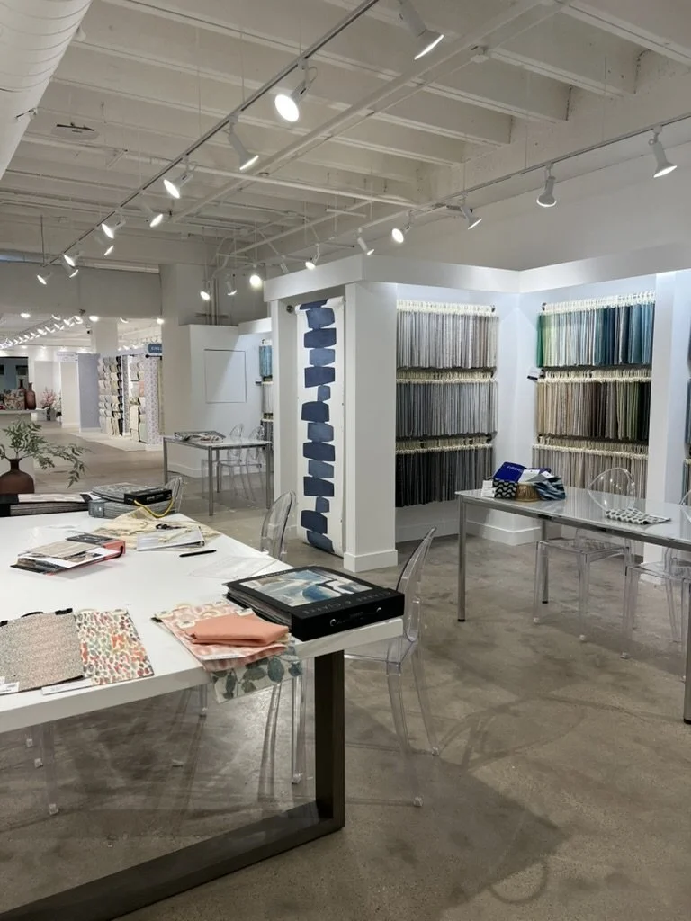 Interior of a showroom displaying fabric and material samples, with a table holding fabric swatches, catalogs, and samples, and walls lined with hanging fabric samples in various colors and textures.