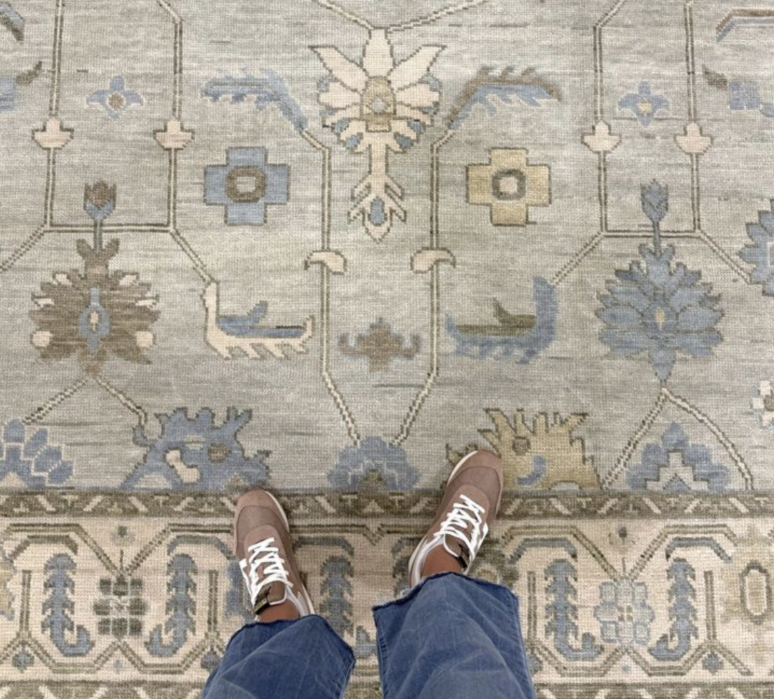 A person standing on an ornate, patterned area rug with beige, blue, and gray hues, wearing brown sneakers and blue jeans.