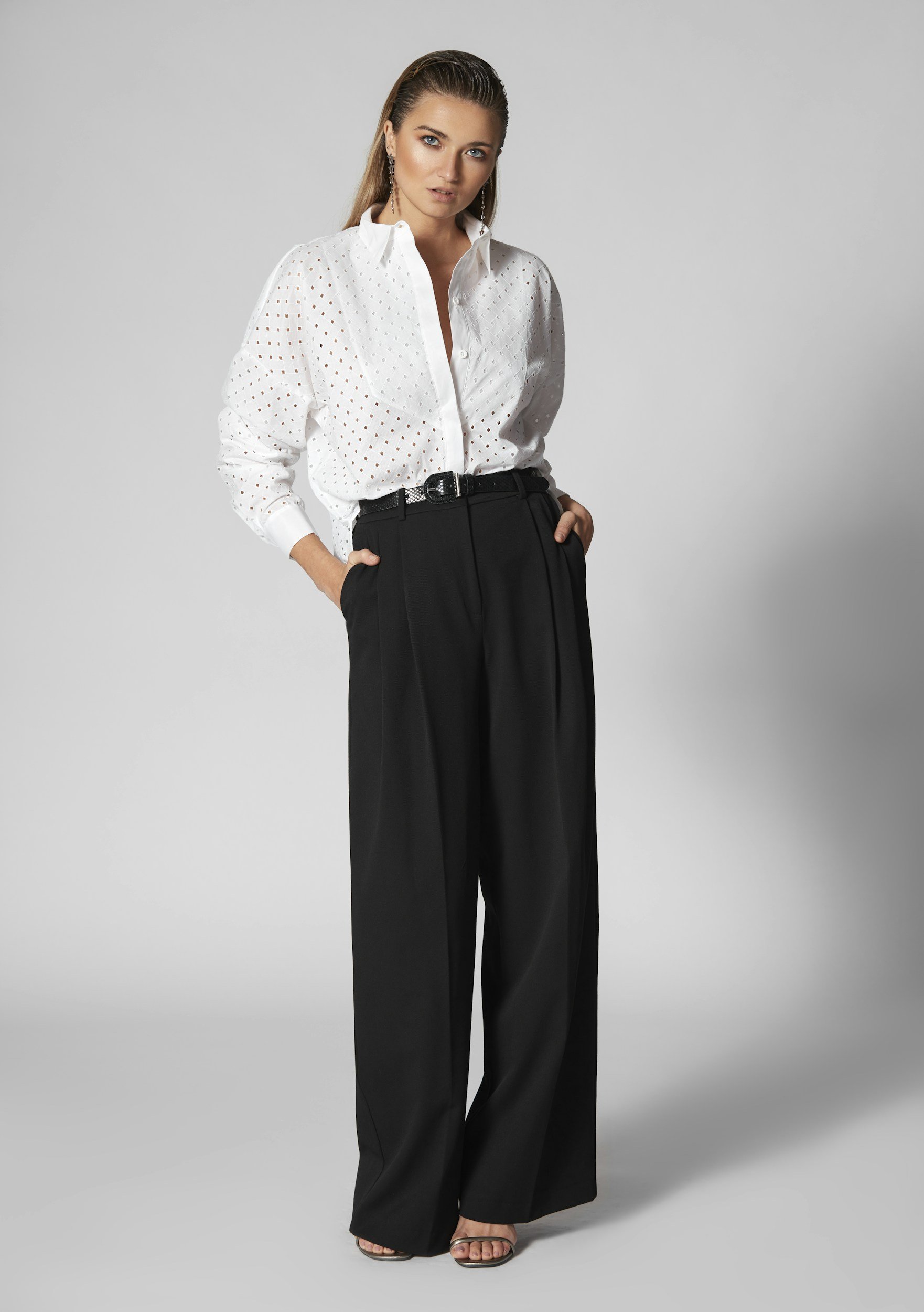 Woman wearing a white perforated long sleeve blouse and black high-waisted wide-leg trousers, standing against a gray background.