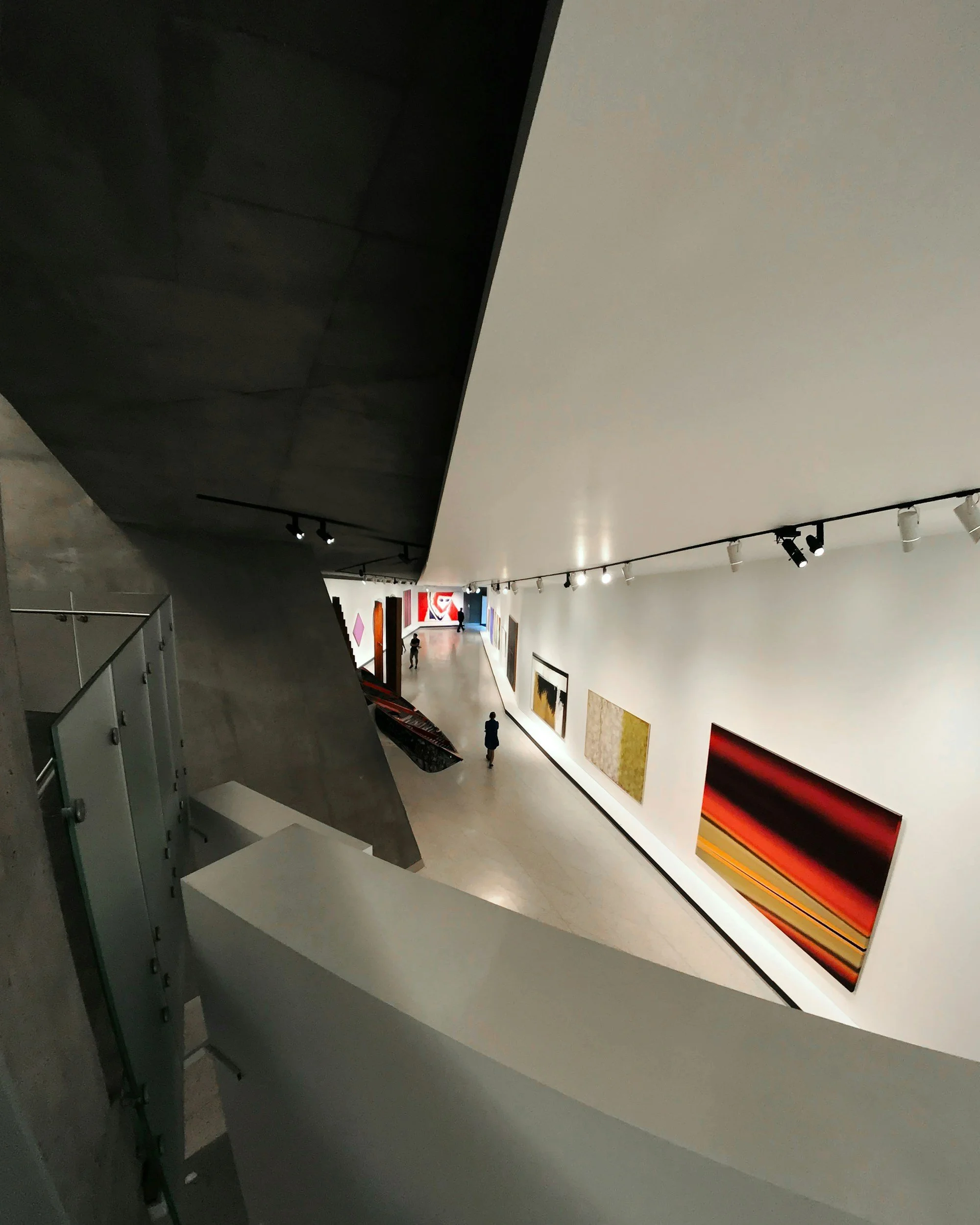 View of an art gallery with colorful abstract paintings on white walls and a few visitors observing the artwork.