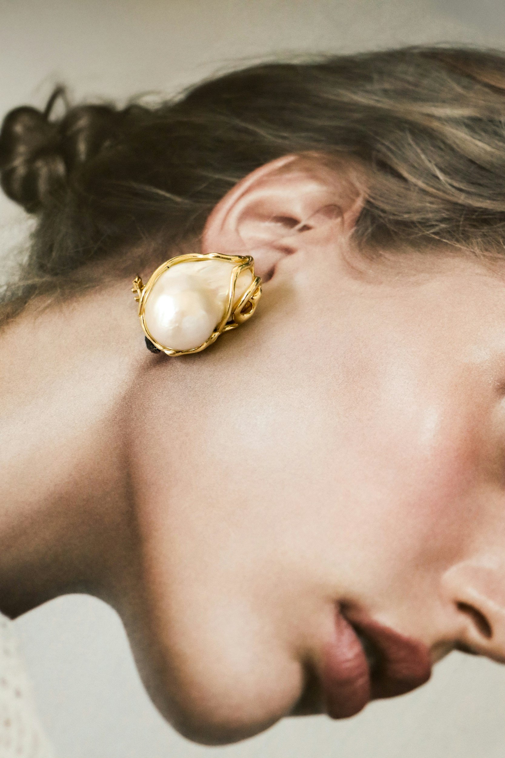 Close-up of a woman wearing a large pearl earring with a gold setting, showing her partially visible face and ear.