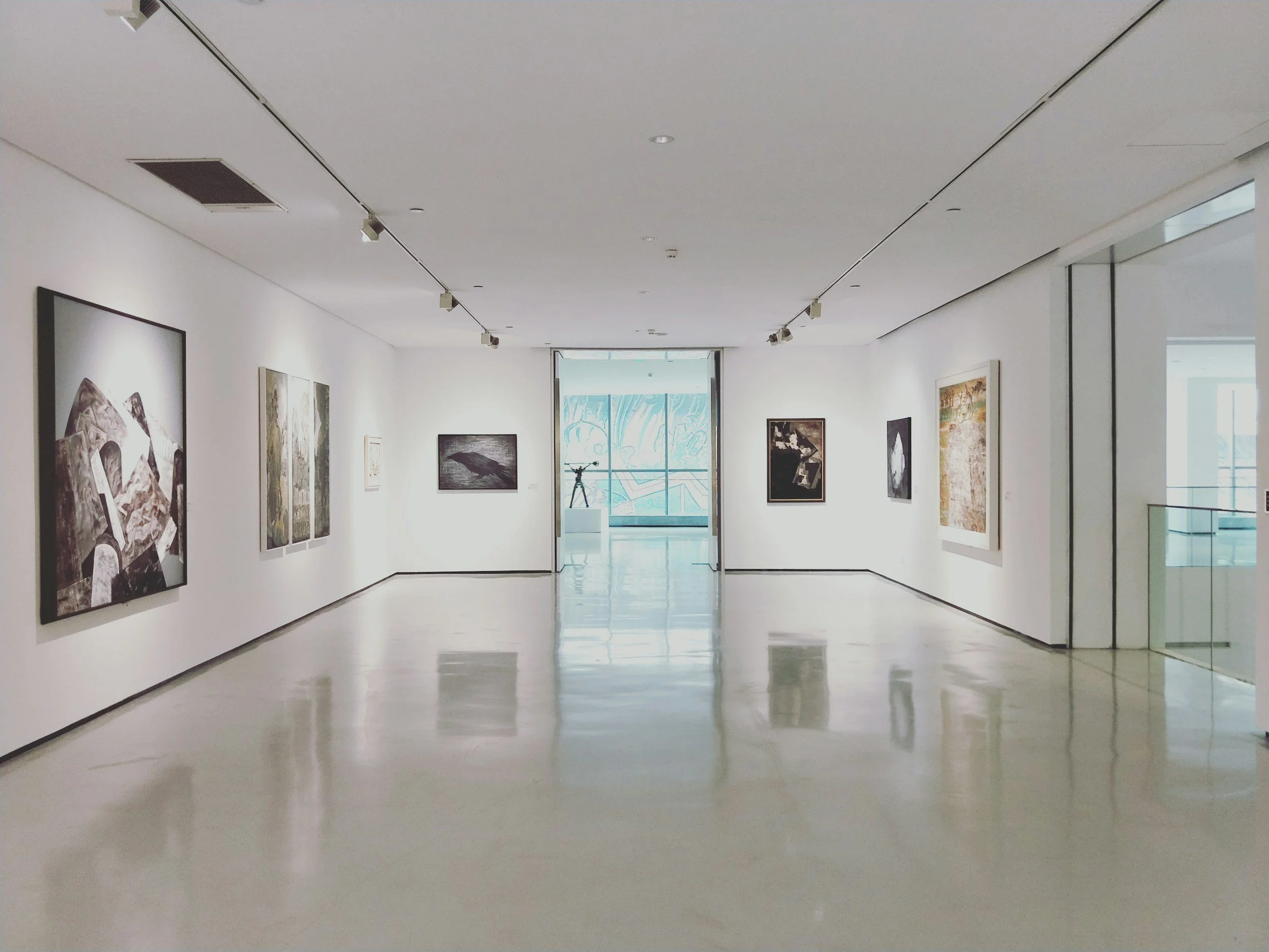 Empty art gallery with paintings on white walls, a telescope on a pedestal near the window, and reflections on a shiny floor.