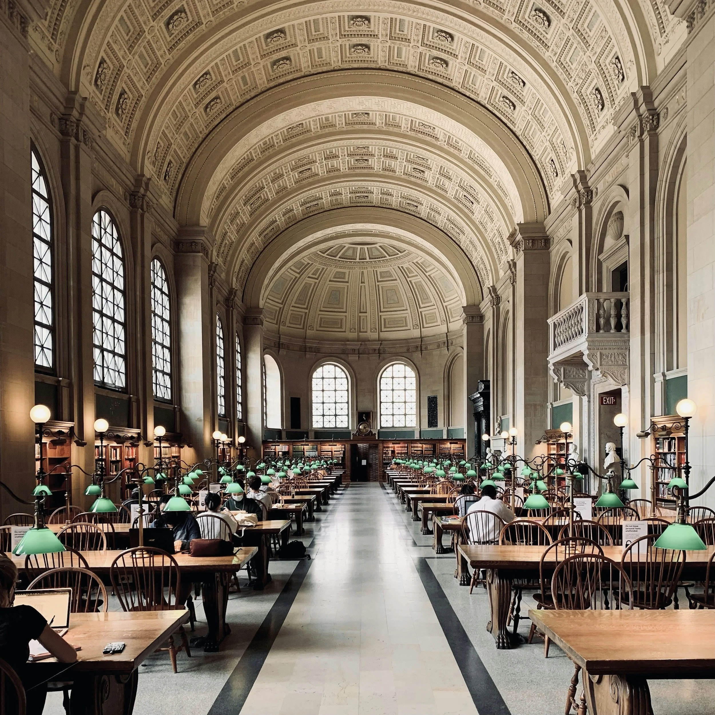 Interior of Boston Public Library with high arched ceilings, tall windows, and rows of wooden tables with green desk lamps. People are seated at the tables reading or working.