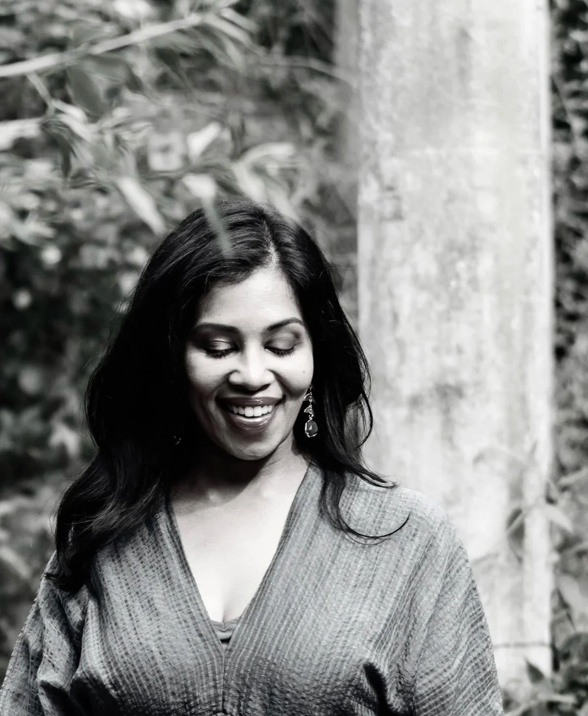 A woman with long dark hair smiling and looking down, wearing earrings and a patterned top, outdoors with foliage in the background.