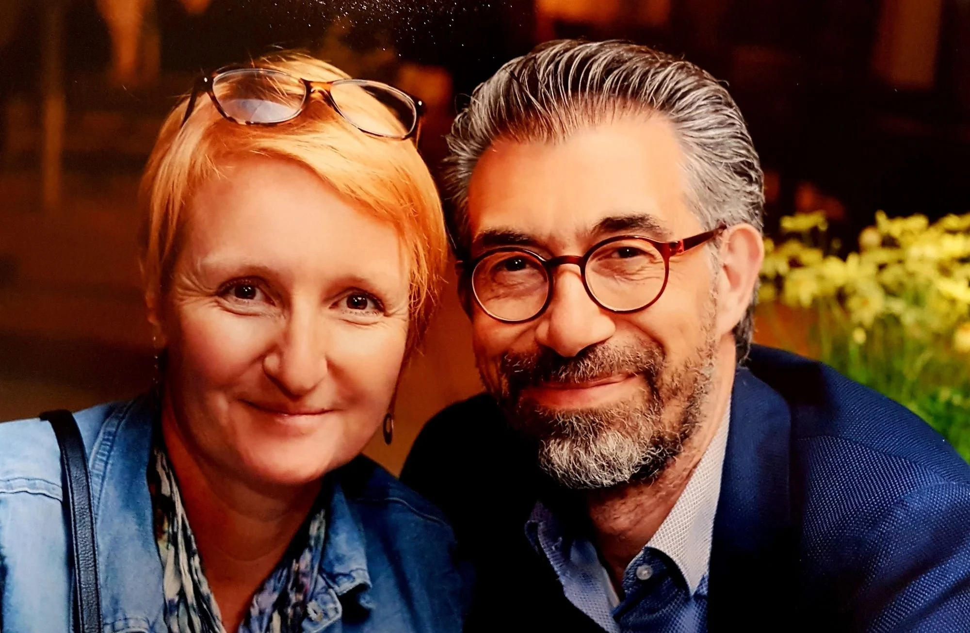 Un couple souriant, la femme a des cheveux rouges courts et porte des lunettes sur la tête, l'homme a une barbe grise, porte des lunettes rondes et un costume, ils posent pour une photo en intérieur avec un décor flou en arrière-plan.