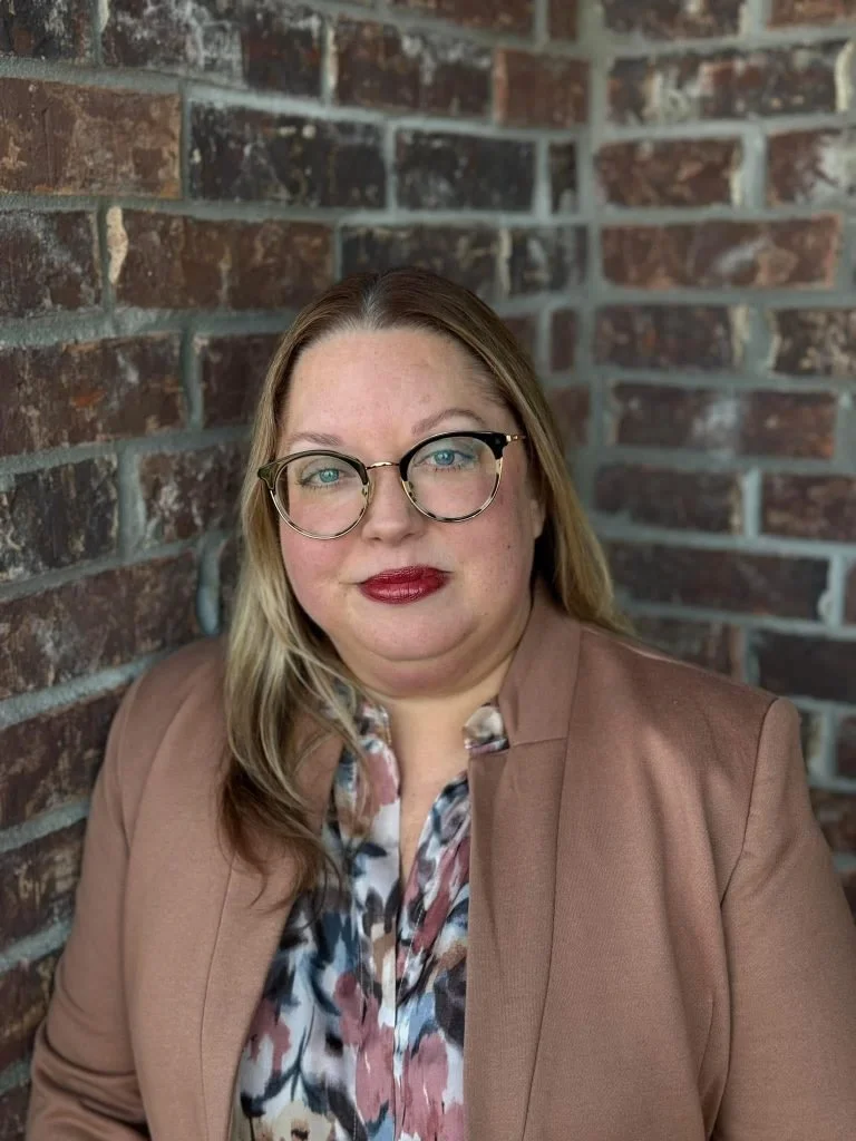 A woman with blonde hair, glasses, and red lipstick, wearing a floral blouse and a tan blazer, standing against a brick wall.