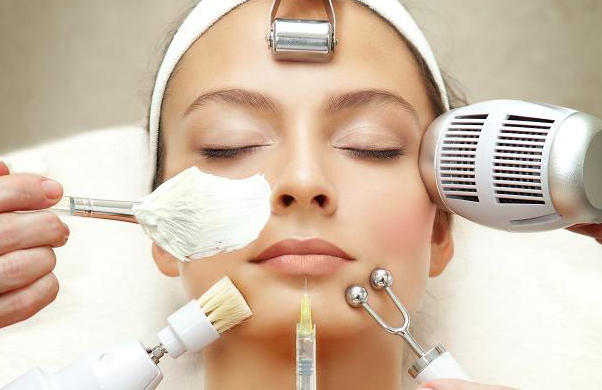 Woman receiving a facial treatment with various skincare tools and a hot towel on her forehead.