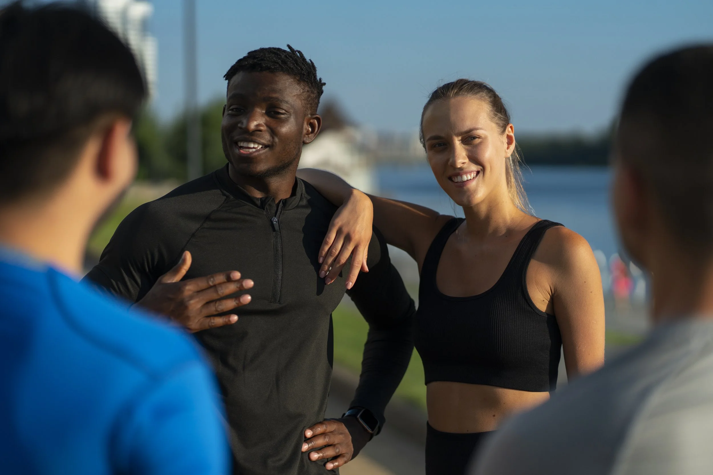 Group of four diverse young adults chatting outdoors near water, dressed in athletic clothing.