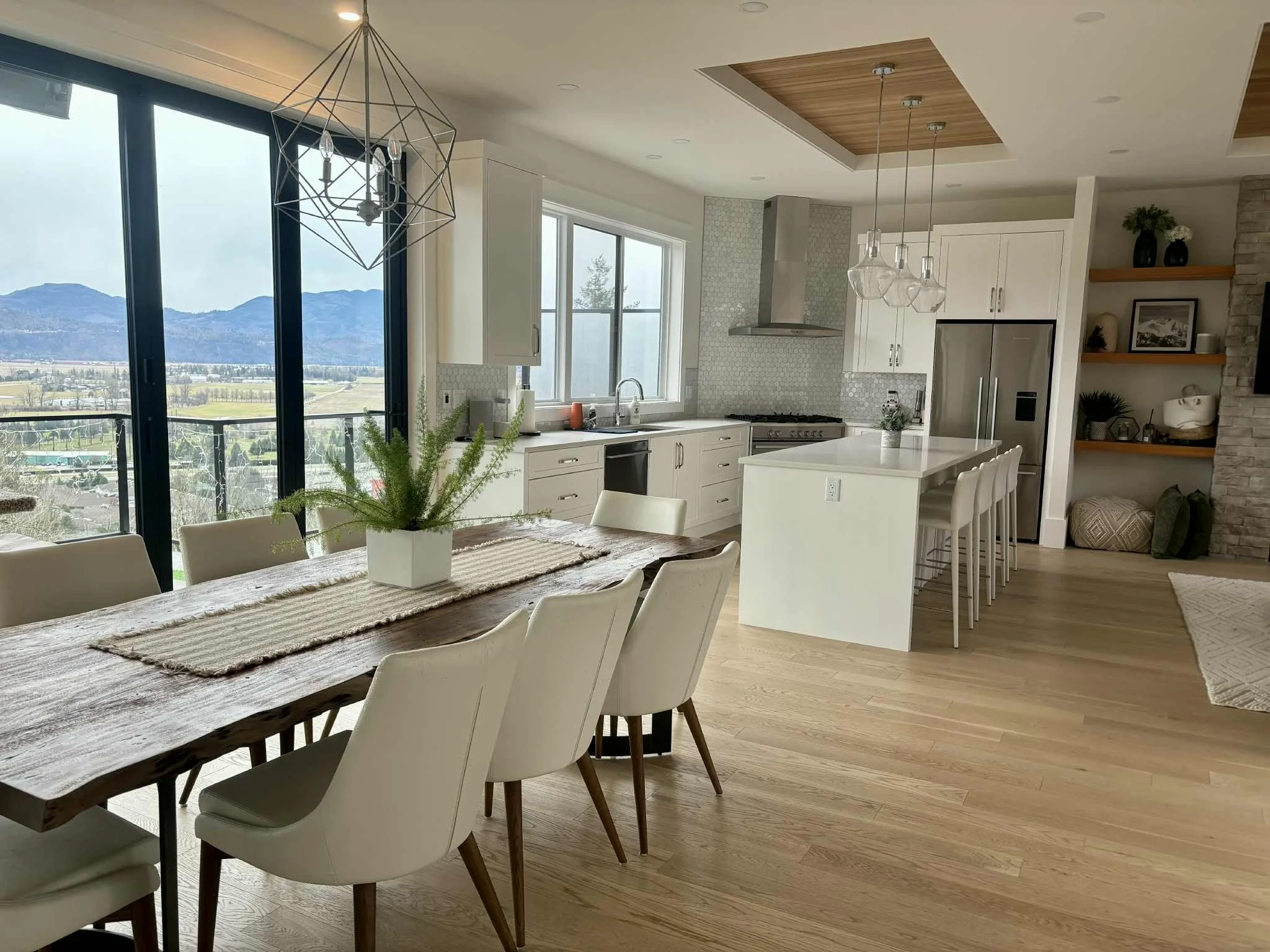 Open-concept kitchen and dining area with mountain view, white cabinets, wooden dining table with white chairs, kitchen island, and large windows