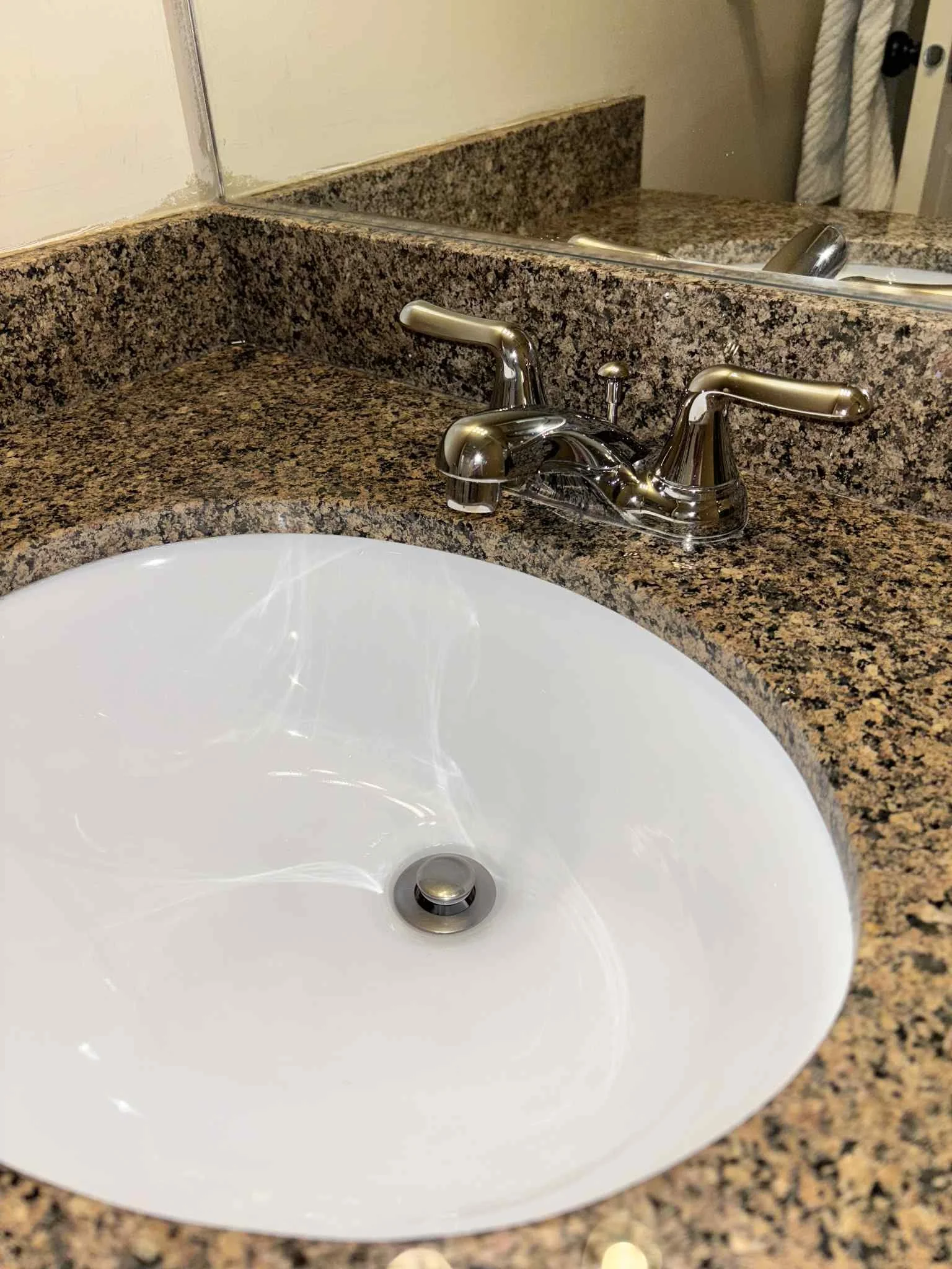 Close-up of a bathroom sink with a granite countertop and a silver faucet, with a mirror reflecting the faucet and part of the wall.