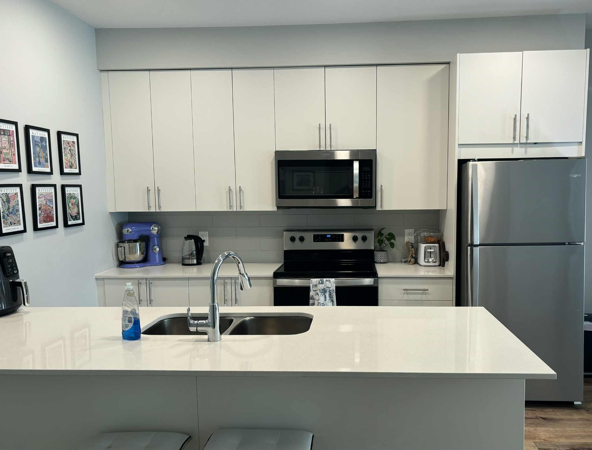 Modern kitchen with white cabinets, stainless steel refrigerator, microwave, stove, and a white countertop with a sink, blue coffee maker, and wall art.
